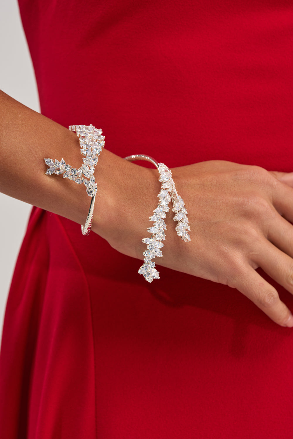 Wearing a bright red dress, the person showcases two LIFE OF THE PARTY BRACELETs—silver, sparkling crystal pieces adorned with clear gemstones arranged like delicate branches—on their wrist and hand.