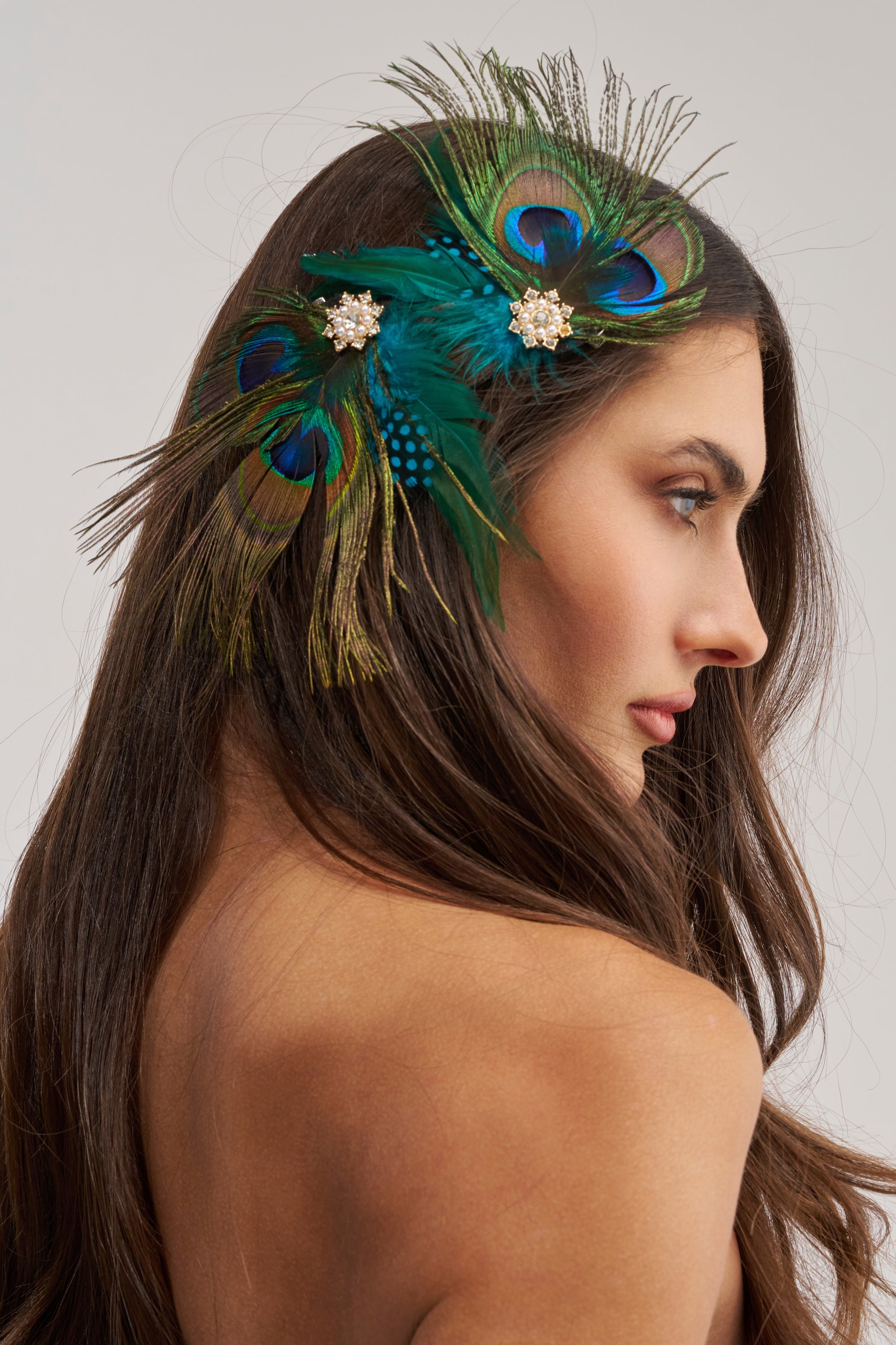 A woman with long brown hair wears the SHOW OFF HAIR CLIP—a peacock feather accessory with jeweled floral details—above her ear. Shown in profile against a plain background, she highlights this bold festival hair piece.