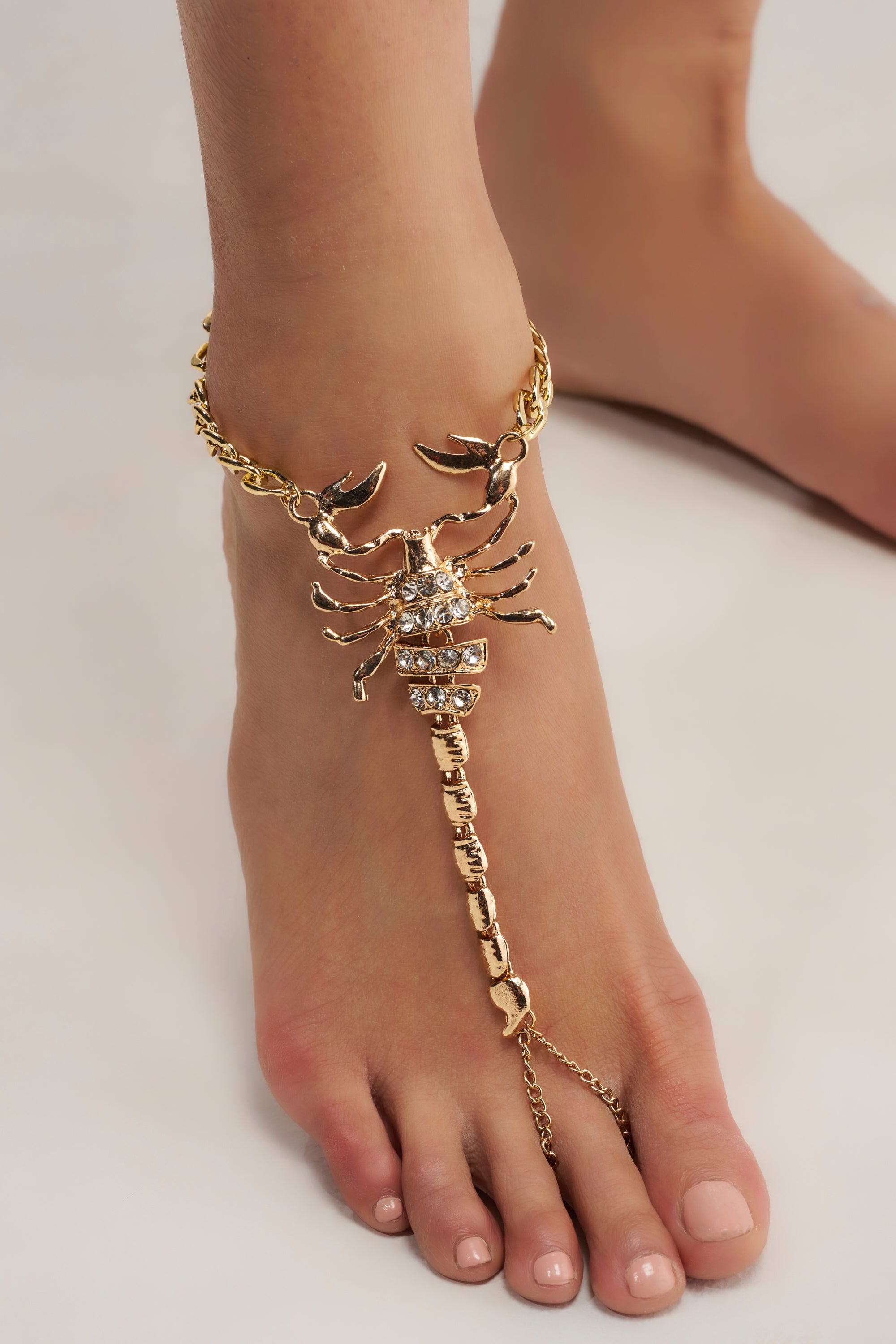 A foot wearing the TOXIC ANKLET, a bold barefoot jewelry piece showcasing a large, rhinestone-studded scorpion charm connected by a chain to a toe ring for an eye-catching look.