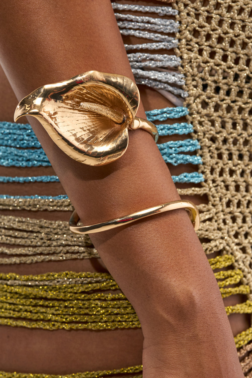 A close-up of a person’s arm wearing two gold bracelets, including the TULIP BRACELET CUFF—a metallic gold arm cuff shaped like a calla lily. They are standing in front of a crocheted garment with horizontal blue, gray, olive, and yellow stripes.
