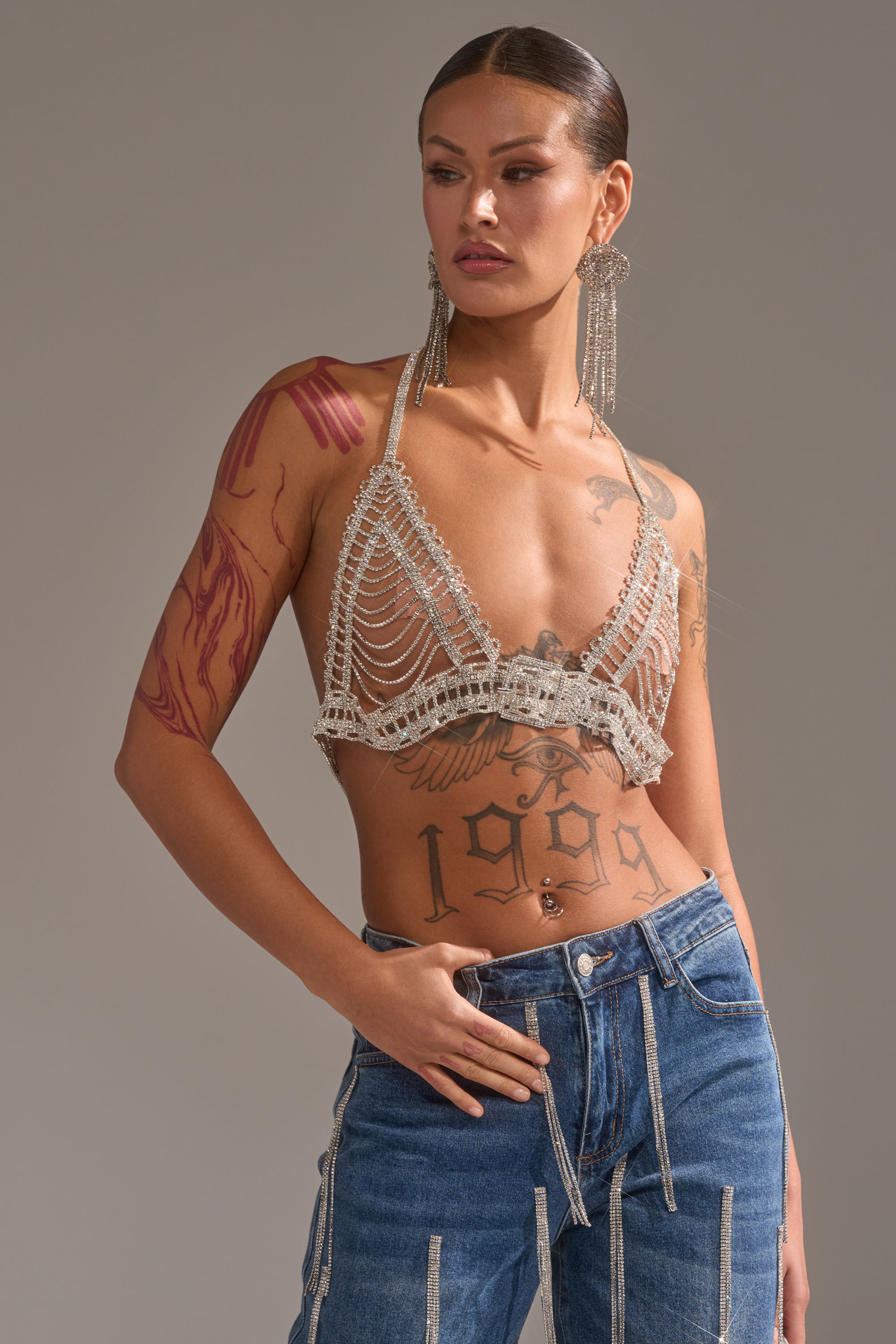 A tattooed woman poses in blue jeans with decorative zippers and the WIND IT UP BODY CHAIN bralette, paired with long dangling earrings. She stands against a neutral background, one hand in her pocket, looking to the side.