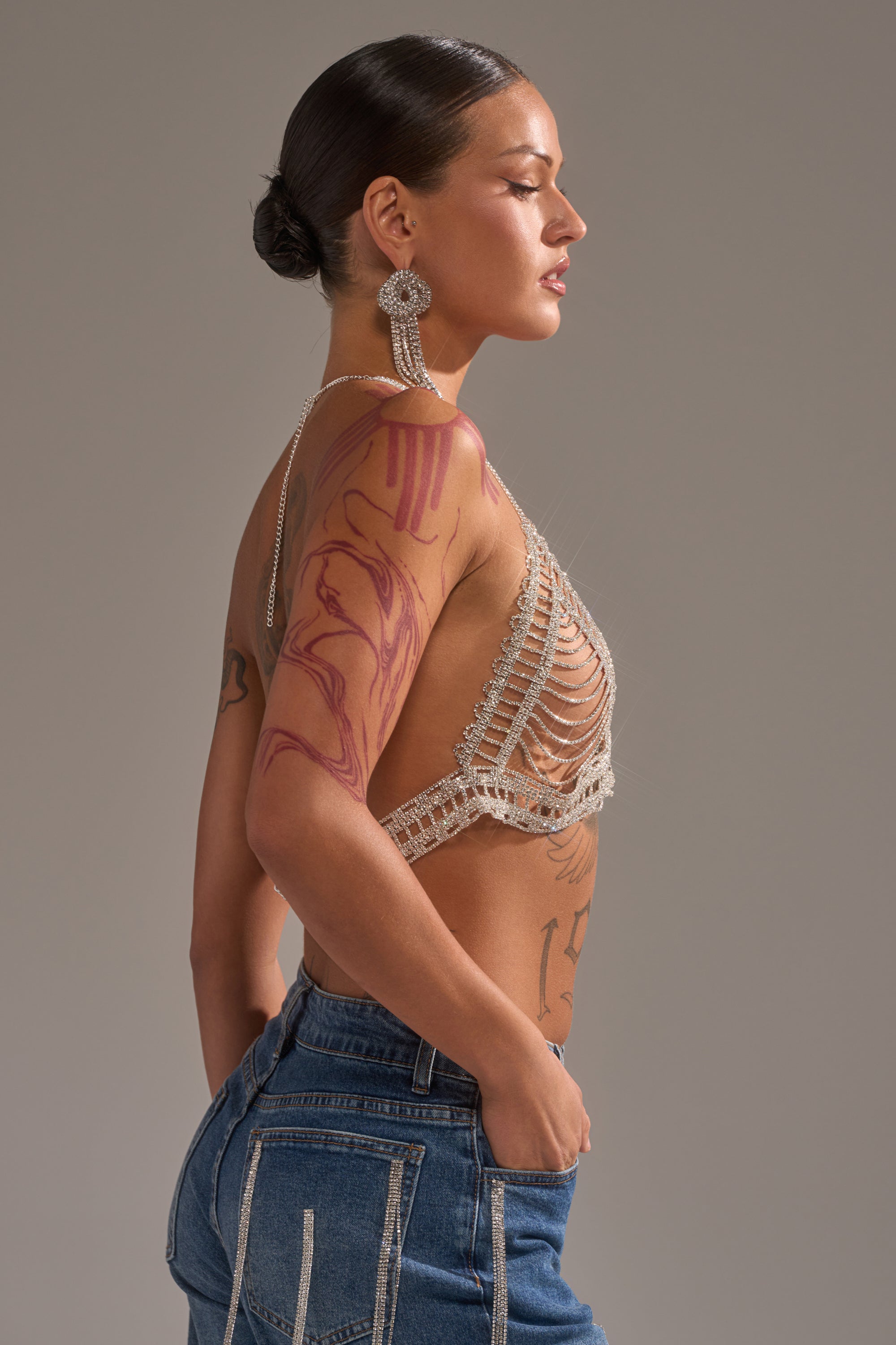 A woman with slicked-back hair and large earrings poses in profile, wearing a beaded top, blue jeans, and the WIND IT UP BODY CHAIN, which adds sparkle to her bold red body art. The background is plain and neutral.