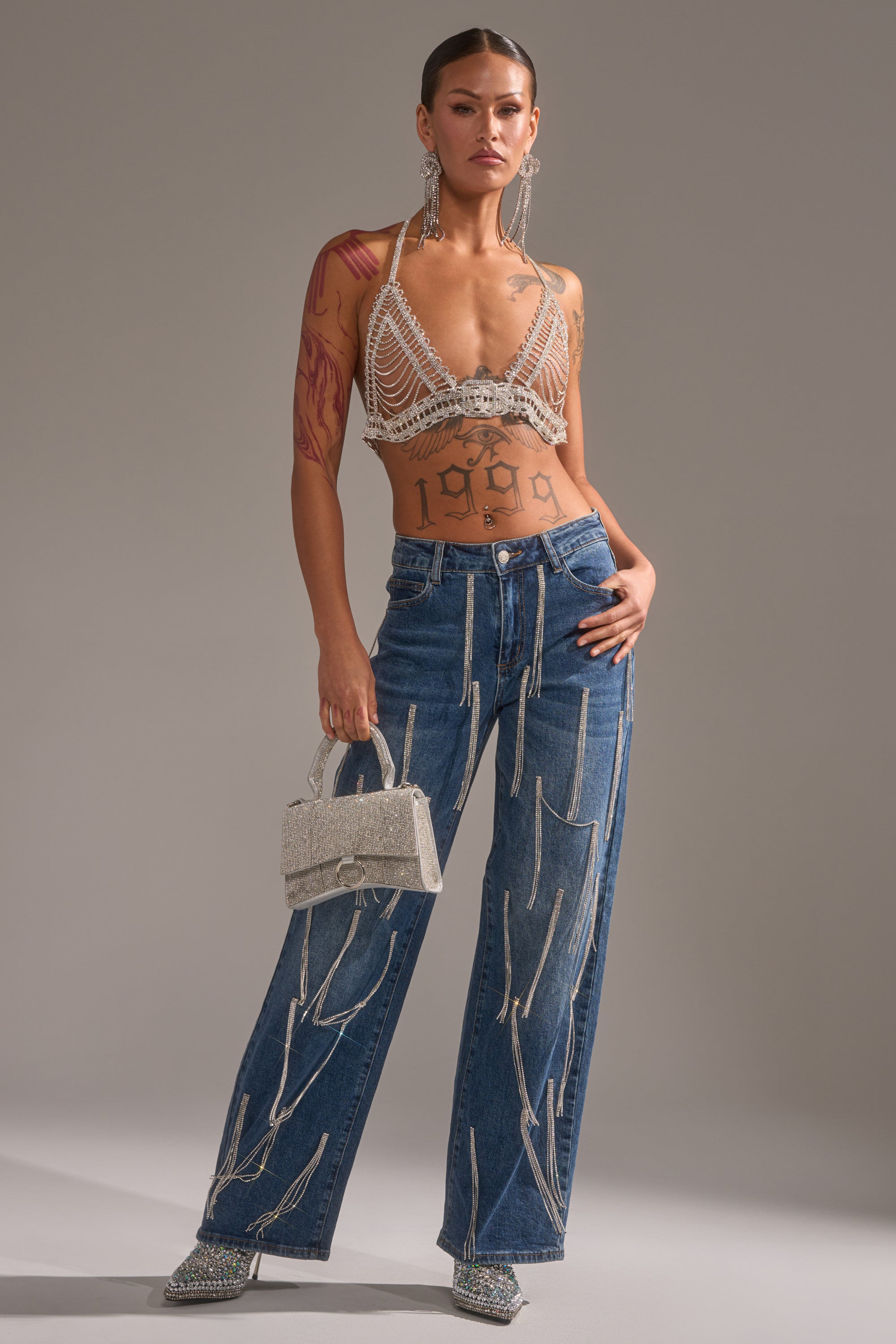 A woman wears the WIND IT UP BODY CHAIN as a festival accessory with a beaded crop top, loose blue jeans with white stitching, silver heels, statement earrings, a sparkly mini handbag, bold makeup, and “1999” on her midriff.