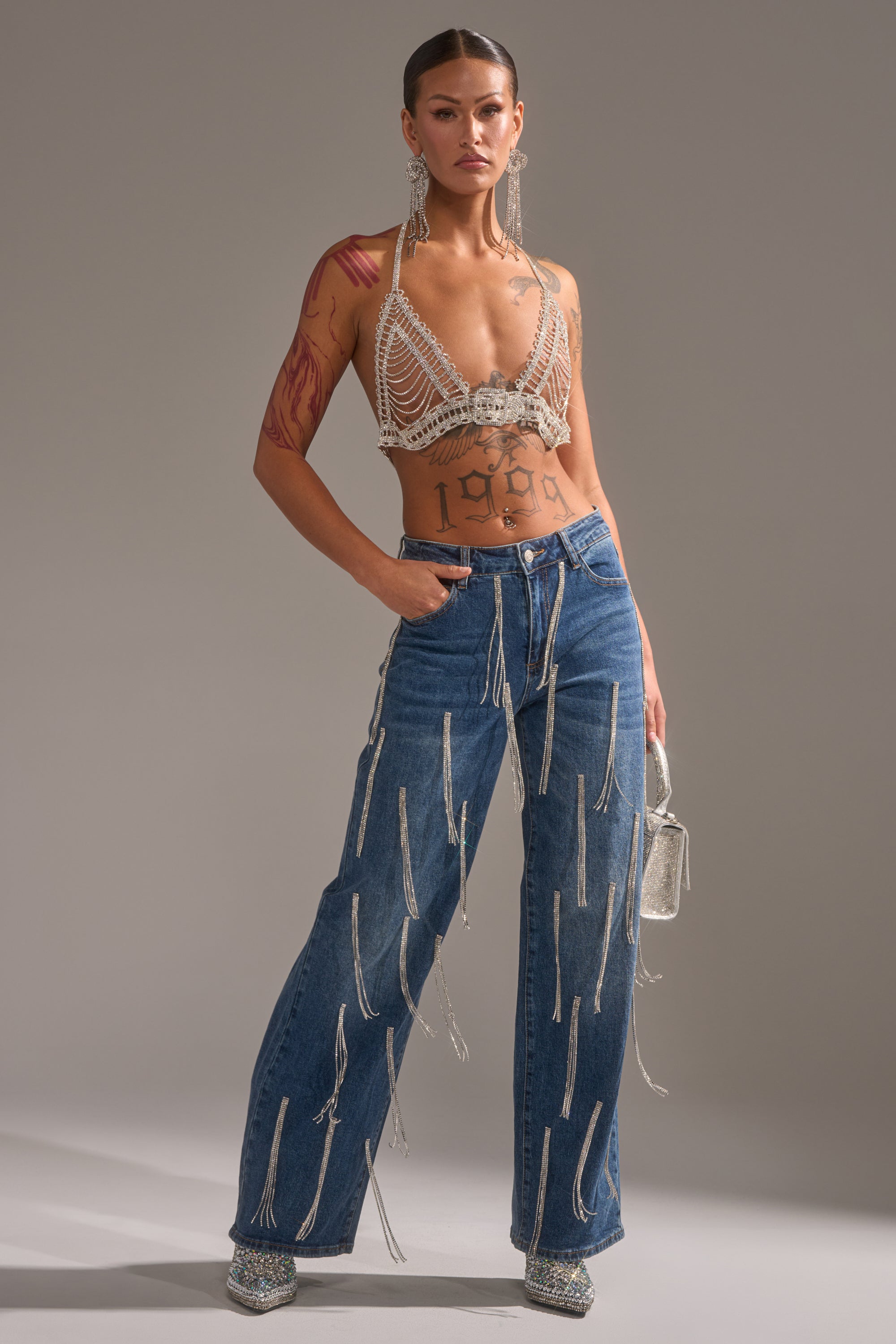 A person poses confidently in wide-leg blue jeans, a beaded bralette, festival jewelry, the WIND IT UP BODY CHAIN, rhinestone earrings, sparkly shoes, and a glittery bag. They showcase bold makeup, tattoos, and a striking expression.