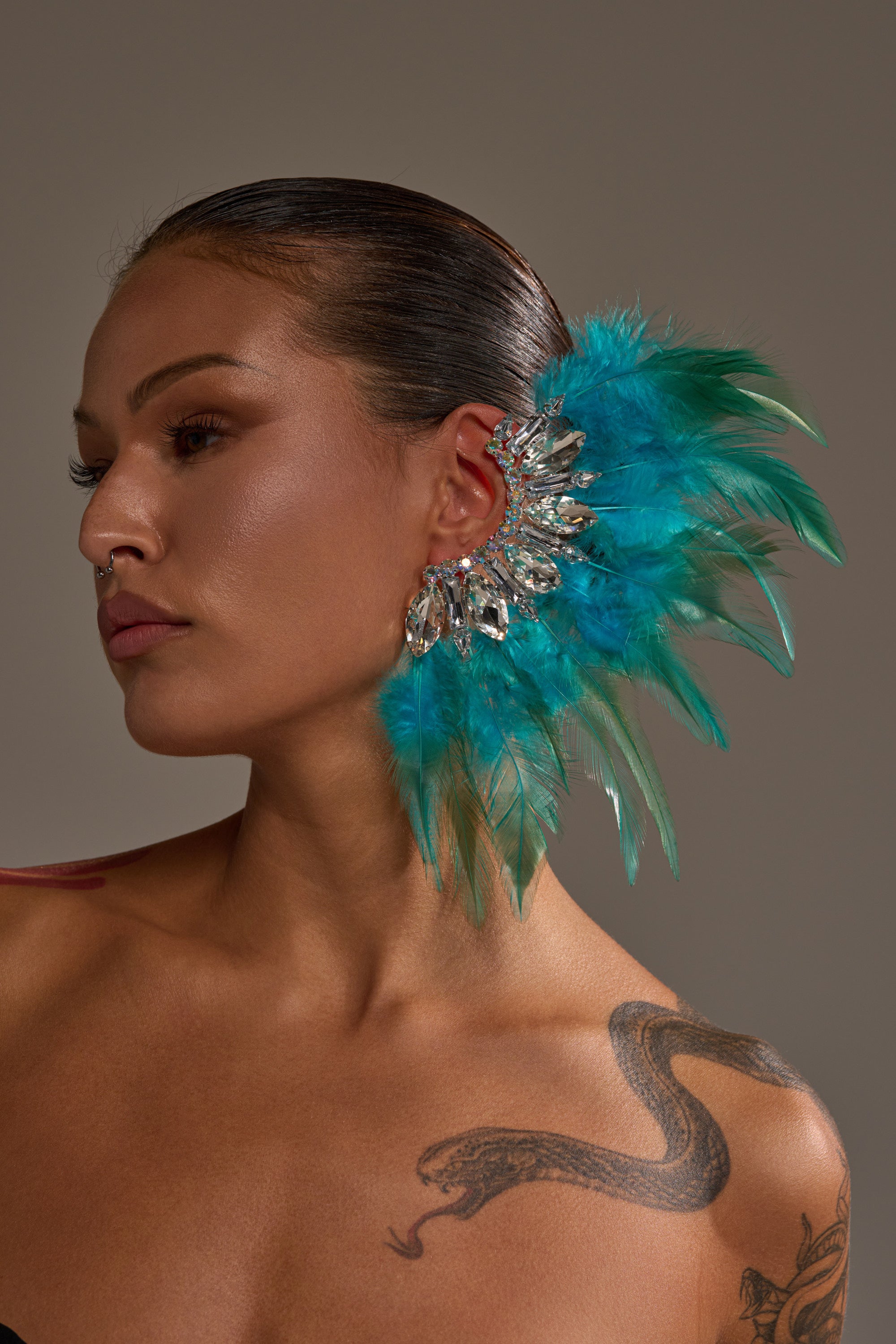 A woman with slicked-back hair showcases the FLY AWAY EARRING, a bold teal and green feathered ear cuff with silver accents. She poses against a neutral background, highlighting her nose ring and snake tattoo on her shoulder.