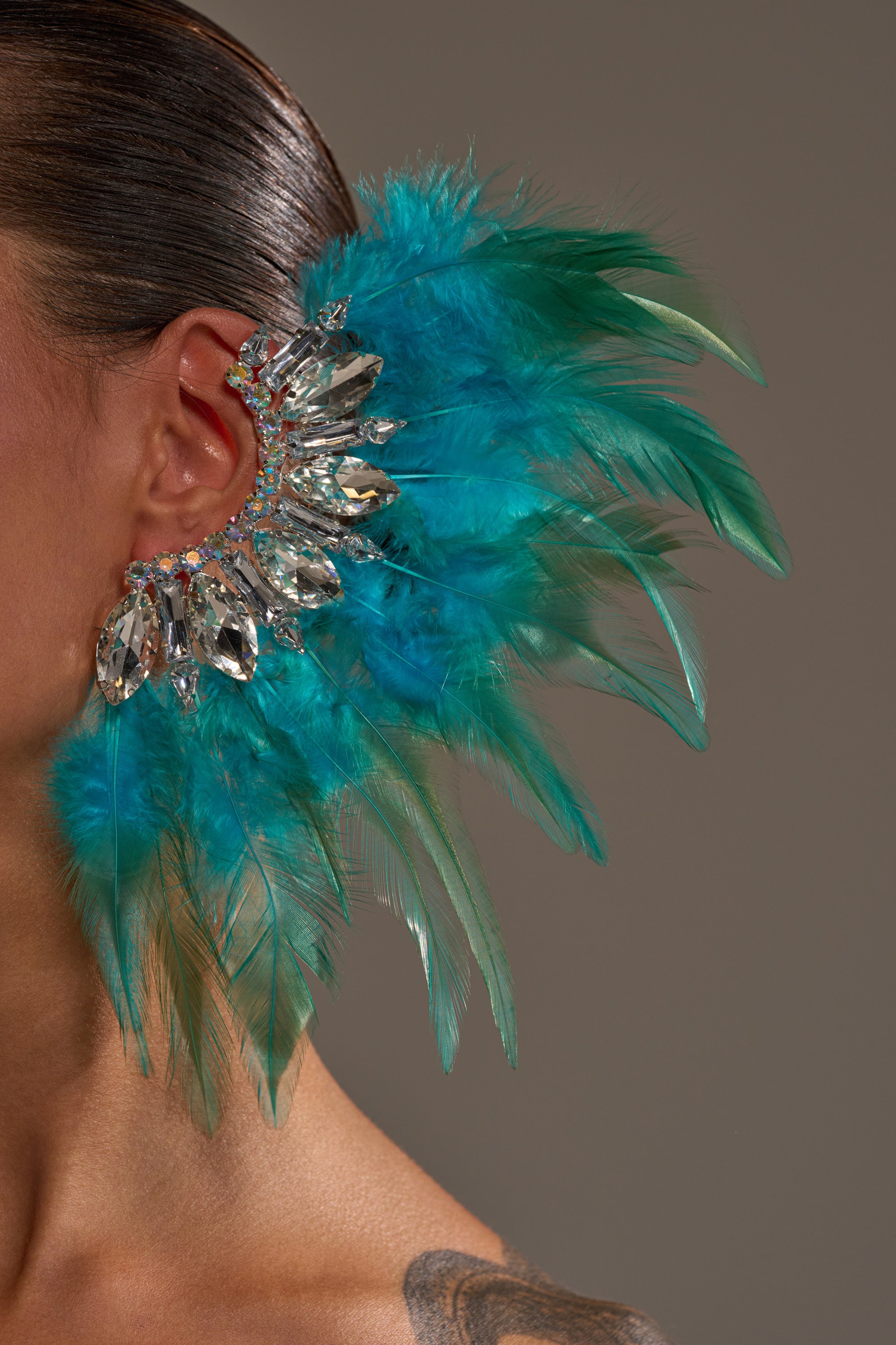Wearing the FLY AWAY EARRING, a striking ear cuff decorated with turquoise feathers and sparkling crystals, this statement accessory covers the ear and extends outward against a plain, neutral background.