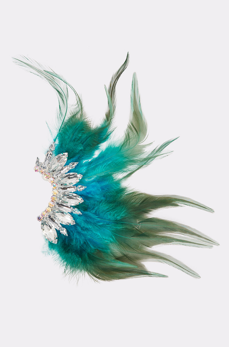 The FLY AWAY EARRING features vibrant teal feathers and green plumes arranged around clear and iridescent gemstones, with the feathers fanned out gracefully to create a striking sense of movement. 1