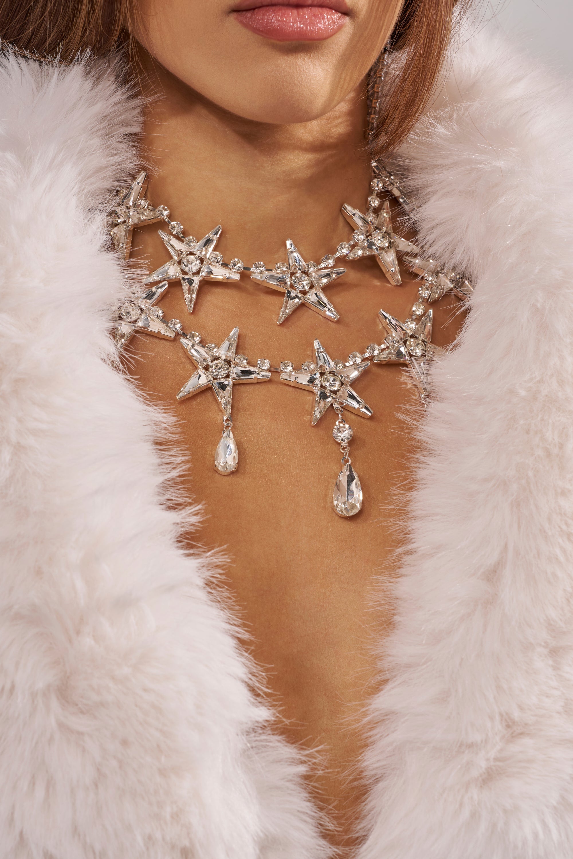A woman in a fluffy white fur coat wears the STARGURL NECKLACE, a celestial statement piece adorned with sparkling star shapes and dangling crystal teardrops, her lips slightly parted. 1
