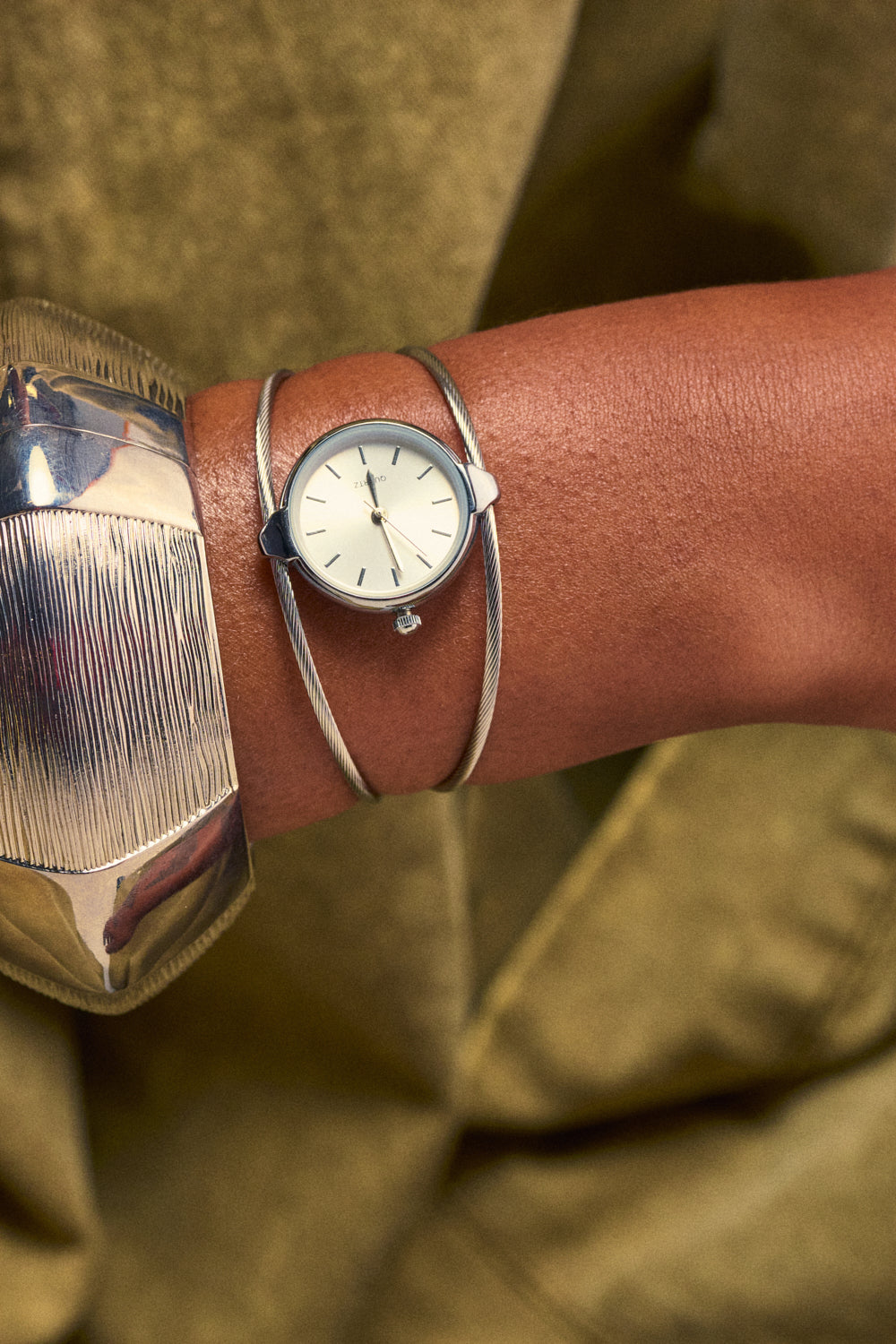 A close-up of a wrist wearing the DOUBLE FUN WATCH BRACELET, featuring a modern silver design with a round, minimalist face and styled elegantly alongside a wide textured silver cuff. The jewelry stack is set against an olive green fabric background.