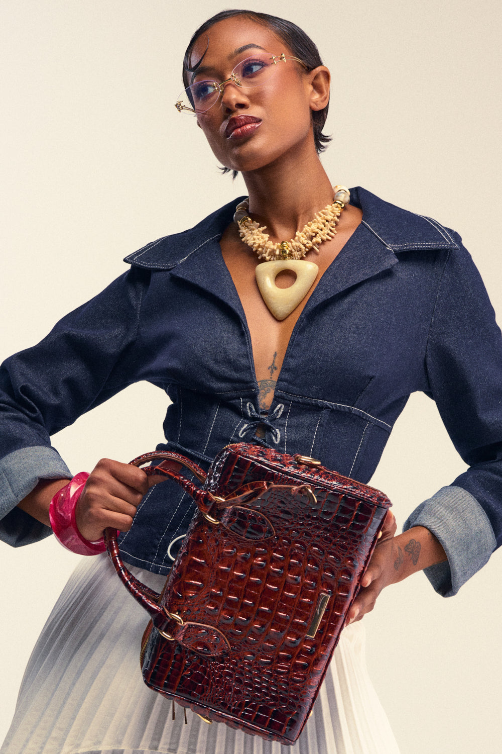 A woman in a denim jacket and white skirt holds the LETS CROC ABOUT IT PURSE, a brown croc faux leather handbag. She wears pink glasses, bold jewelry—including a chunky necklace and bracelet—and has short hair with tattoos on her hands.