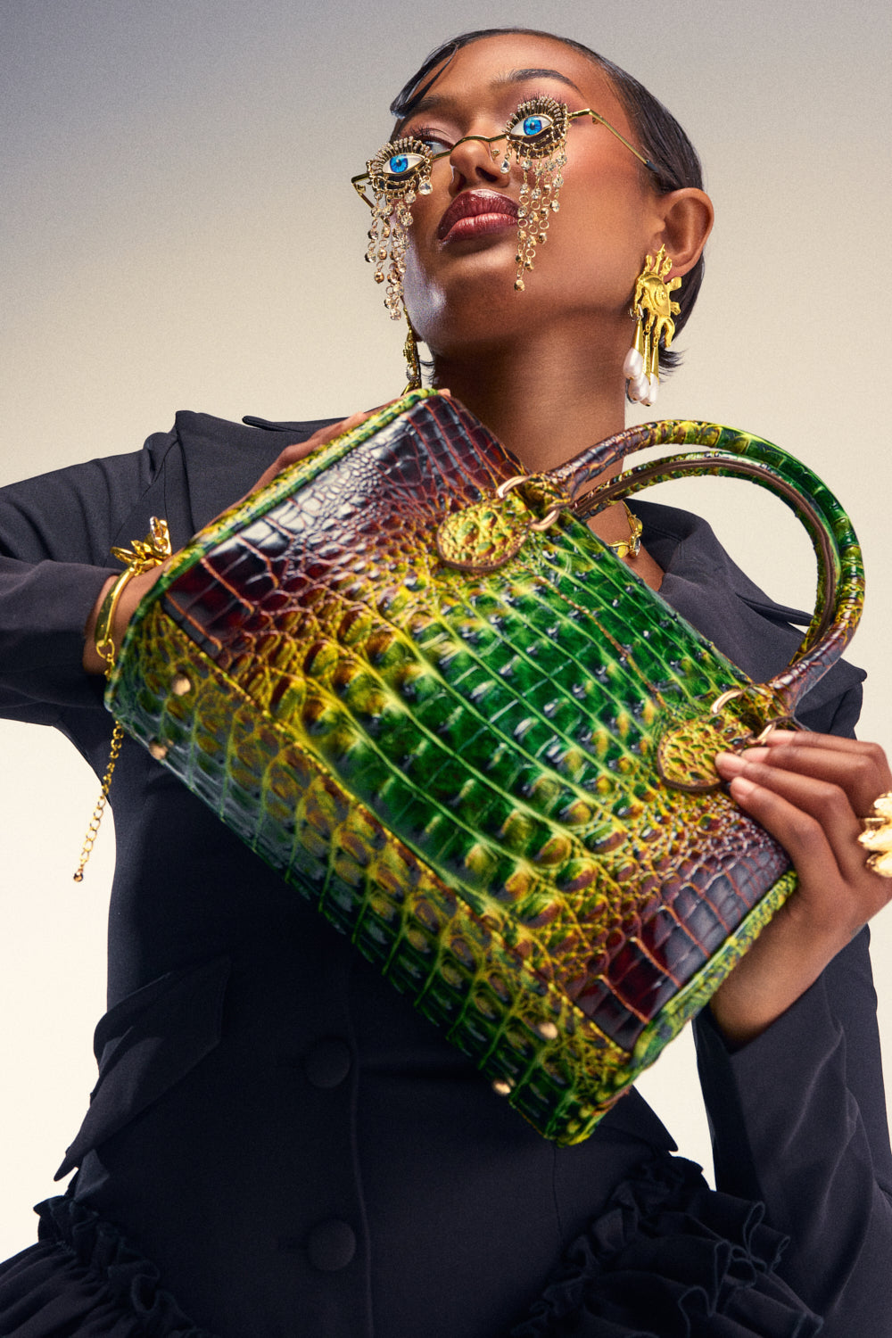 A stylish person dressed in black showcases the RATTLE SNAKE BAG—a large green and yellow faux leather snakeskin purse with streetwear flair. They accessorize with striking gold jewelry, including statement earrings and glasses adorned with dangling chains, all set against a neutral backdrop.