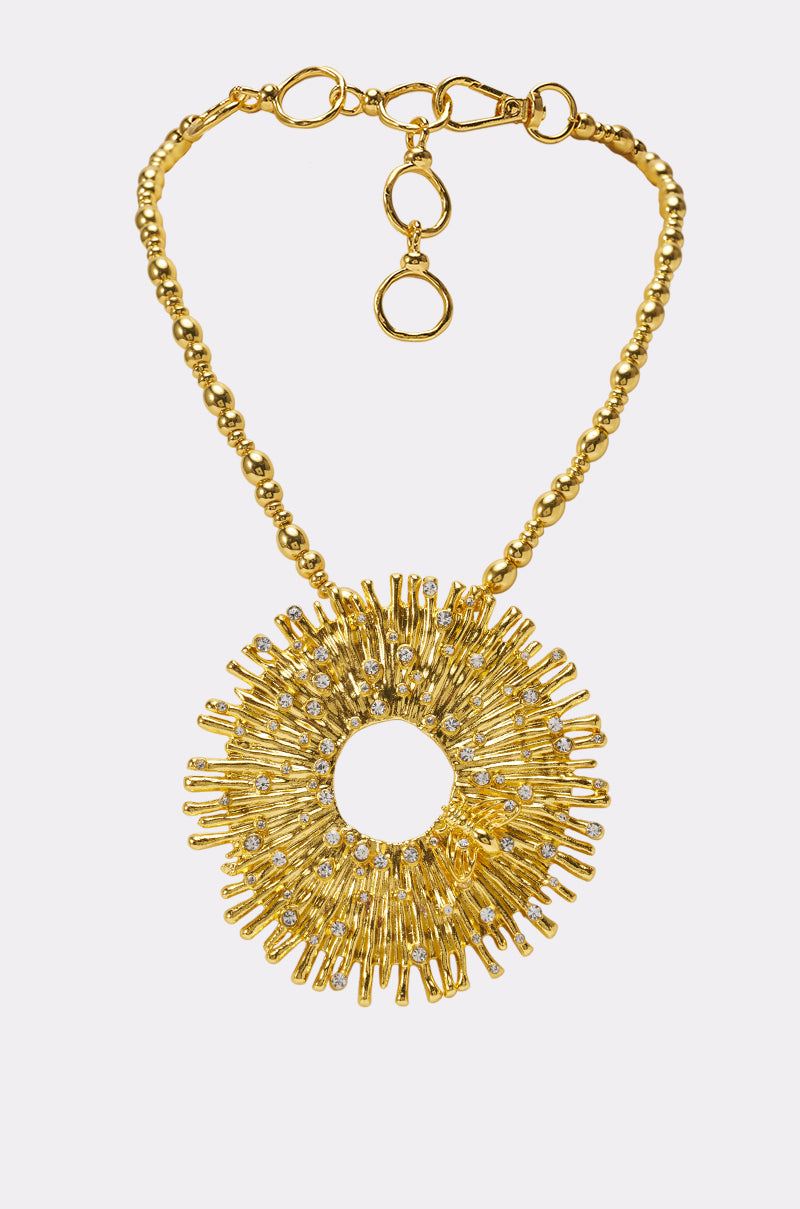 The MATTE GOLD STARBURST NECKLACE features a large, circular sunburst pendant adorned with radiating spikes and sparkling clear stones, all showcased against a white background—an elegant statement piece that brings radiant charm to any look.