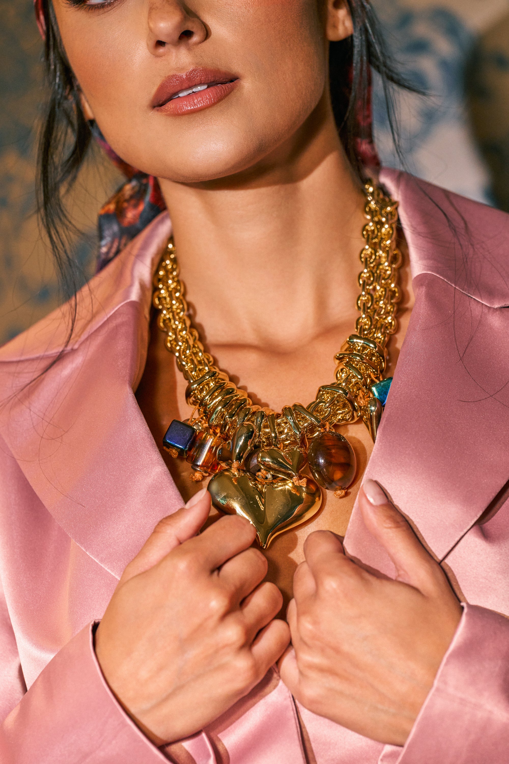 Wearing the MIXED MEDIA HEART STATEMENT NECKLACE—a chunky gold piece adorned with heart and gemstone charms—a woman in a pink satin blazer holds the lapels, creating a striking statement look.