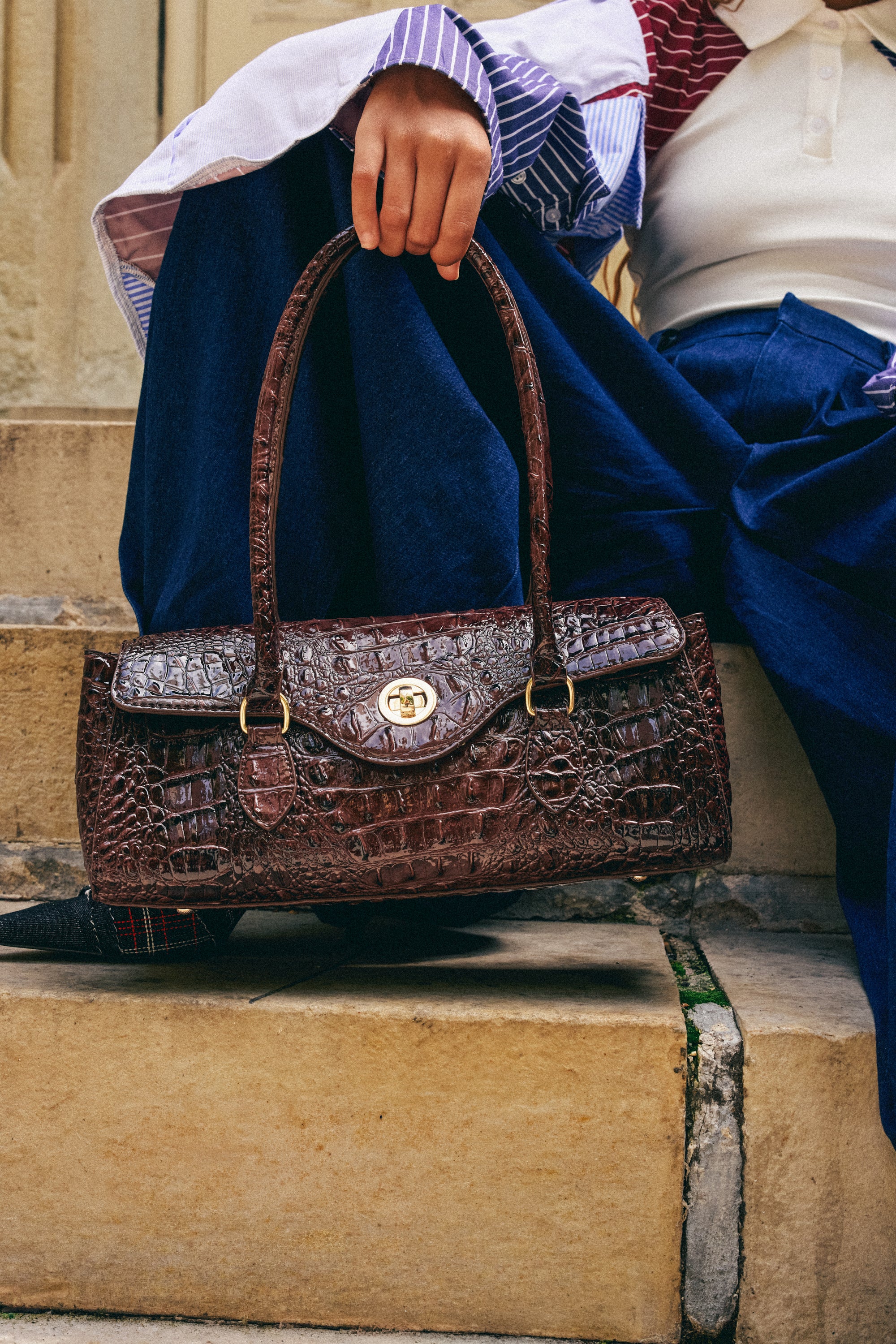 A person sitting on stone steps holds the chic CROCODILE TEARS PURSE with gold hardware, wearing wide-leg blue pants and a striped shirt.
