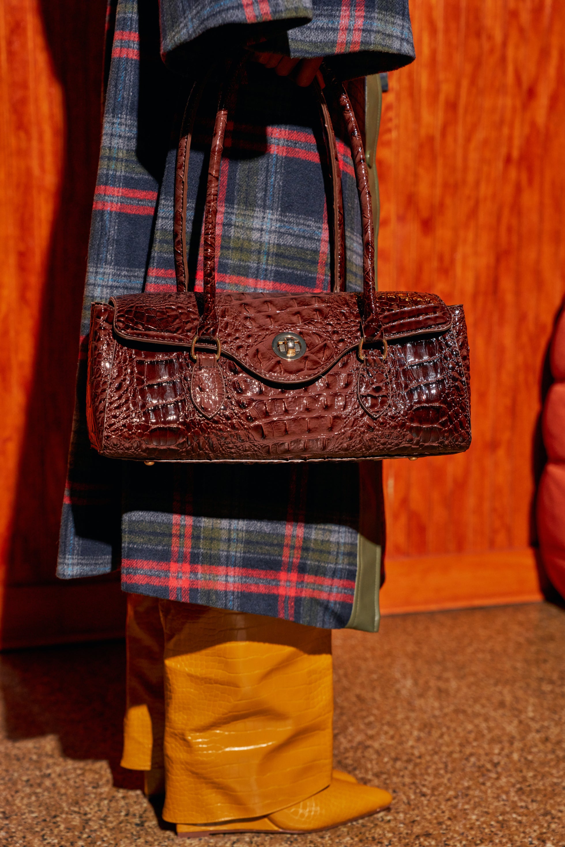 A person wearing a plaid coat and yellow boots carries a rectangular burgundy CROCODILE TEARS PURSE, a croc-embossed barrel bag with a silver turn-lock closure.