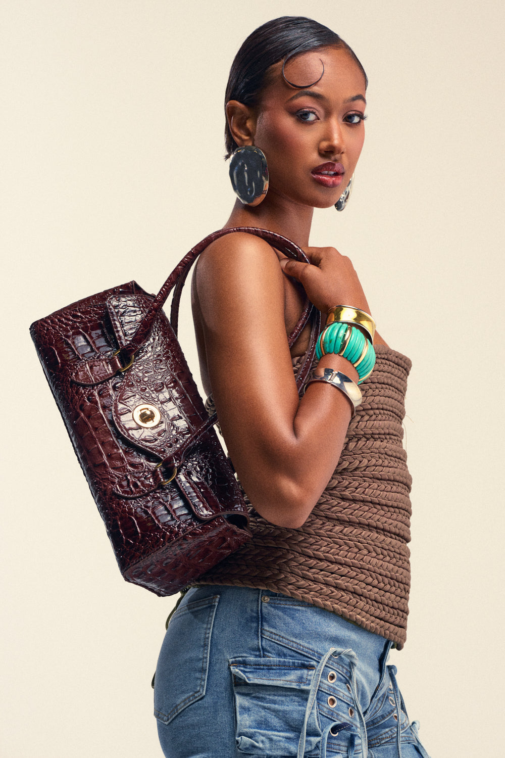 A woman wearing a brown textured top, blue cargo pants, large earrings, colorful bangles, and carrying the chic CROCODILE TEARS PURSE over her shoulder poses against a neutral background.