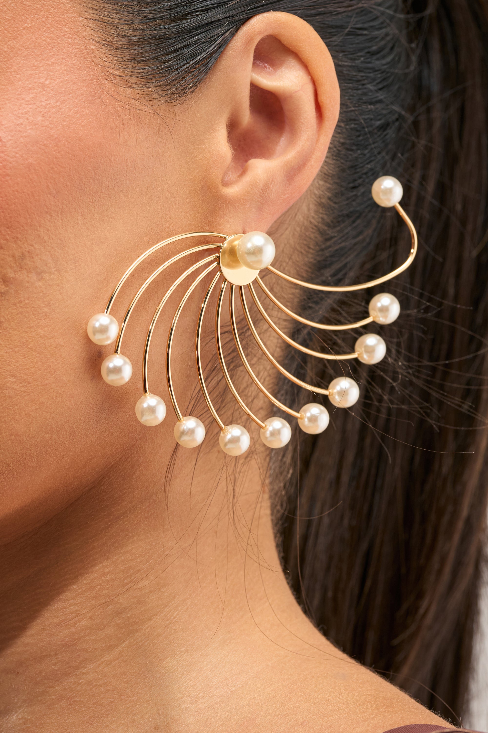 A close-up of a woman's ear wearing the PEARL EAR HUGGERS, large gold fan-shaped earrings adorned with multiple pearl accents on each arc, styled against her dark, pulled-back hair; this striking design evokes modern bridal earrings and doubles as an elegant statement ear cuff.
