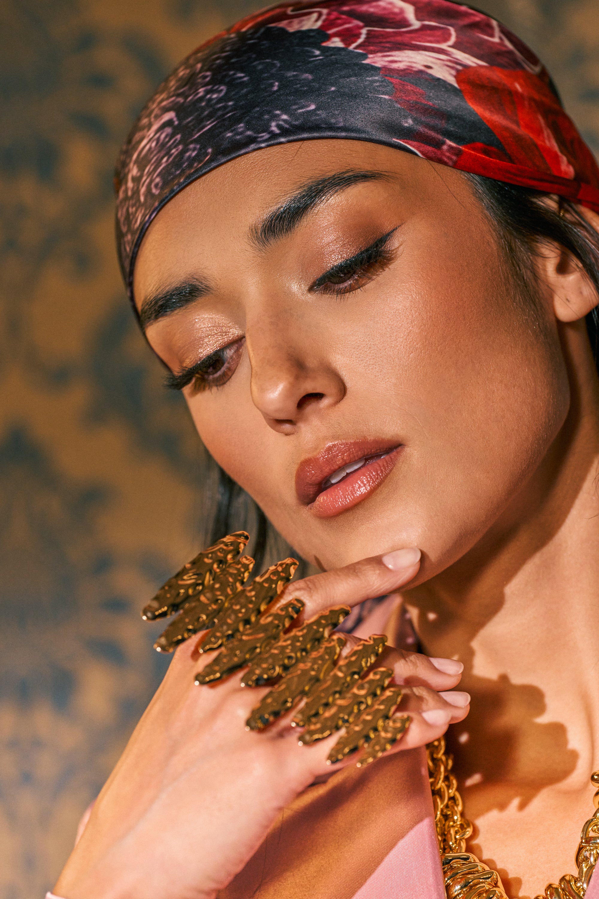 A woman wearing a patterned headscarf poses thoughtfully, her hand touching her chin and her eyes cast downward. She showcases natural makeup, a gold necklace, and the HEAT WAVES RING AND BRACELET SET IN GOLD—an elegant statement ensemble that enhances her look with its striking ring and cuff bracelet combination.