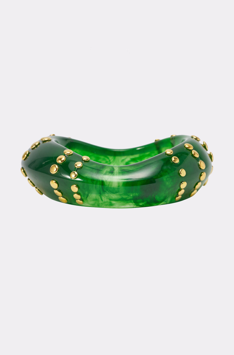 The COOL GIRL STUDDED BANGLE IN GREEN features a chunky, glossy acrylic construction in a vibrant green hue with a wavy silhouette, embellished with clusters of small round gold studs for a bold statement look. This eye-catching gold studded bangle is shown against a plain white background.