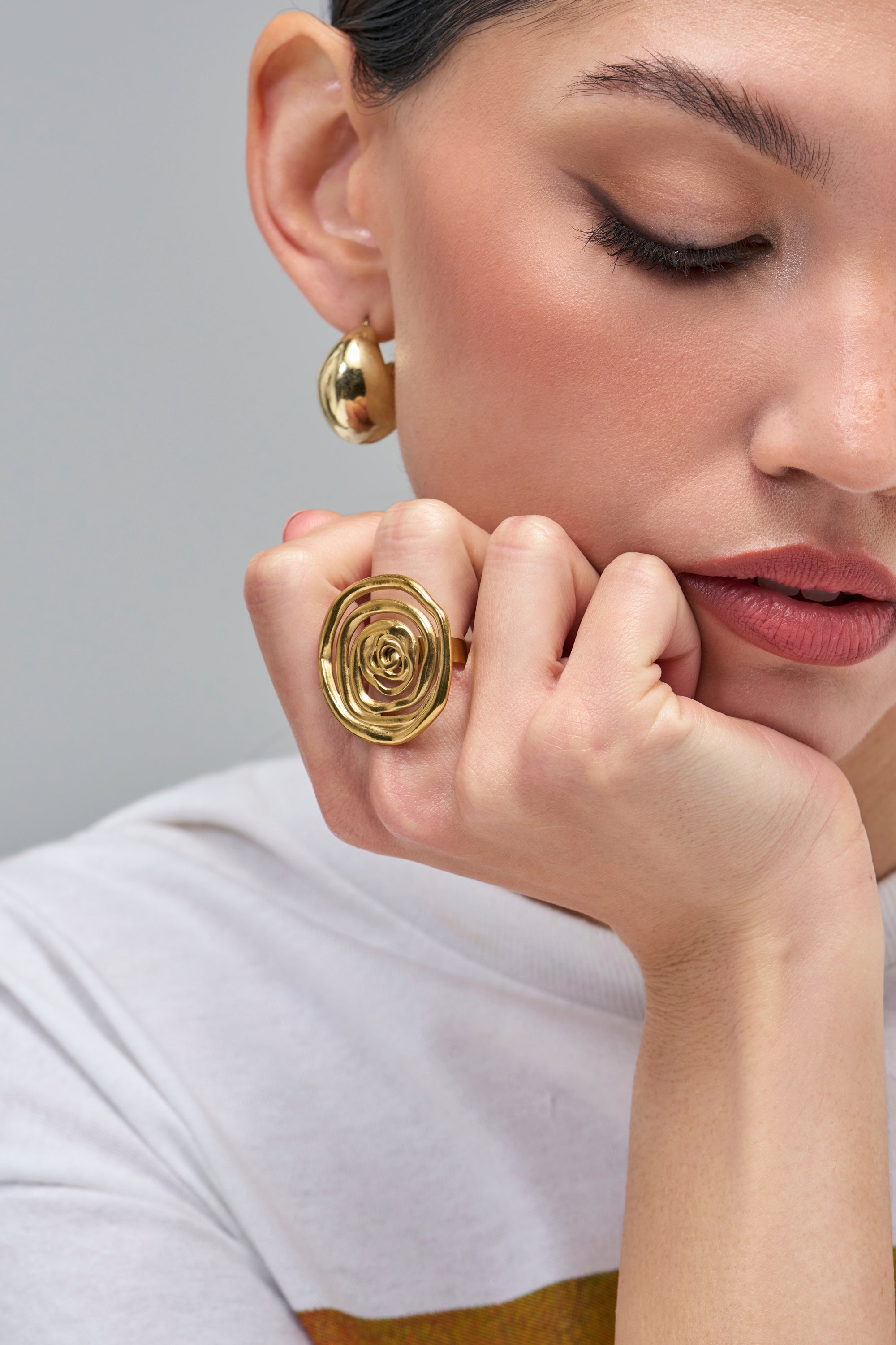 A gold WHIRLWIND RING with a swirling, spiral design and an adjustable band worn on a finger resting against a cheek.