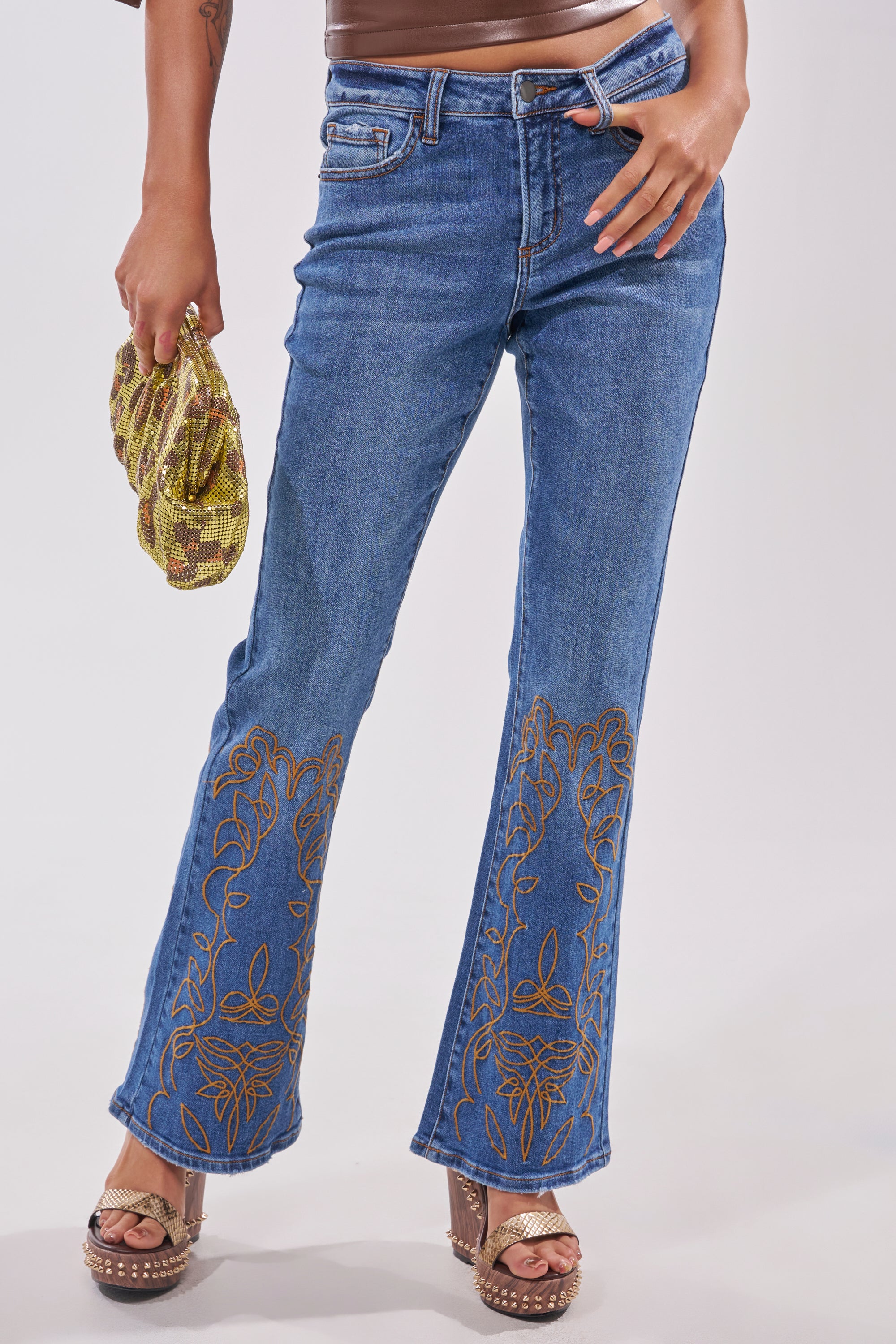 A person wears RODEO BABE SKINNY FLARED DENIM with gold embroidery at the hems, brown studded platform sandals, and holds a gold sequin clutch against a plain light-colored background. 0