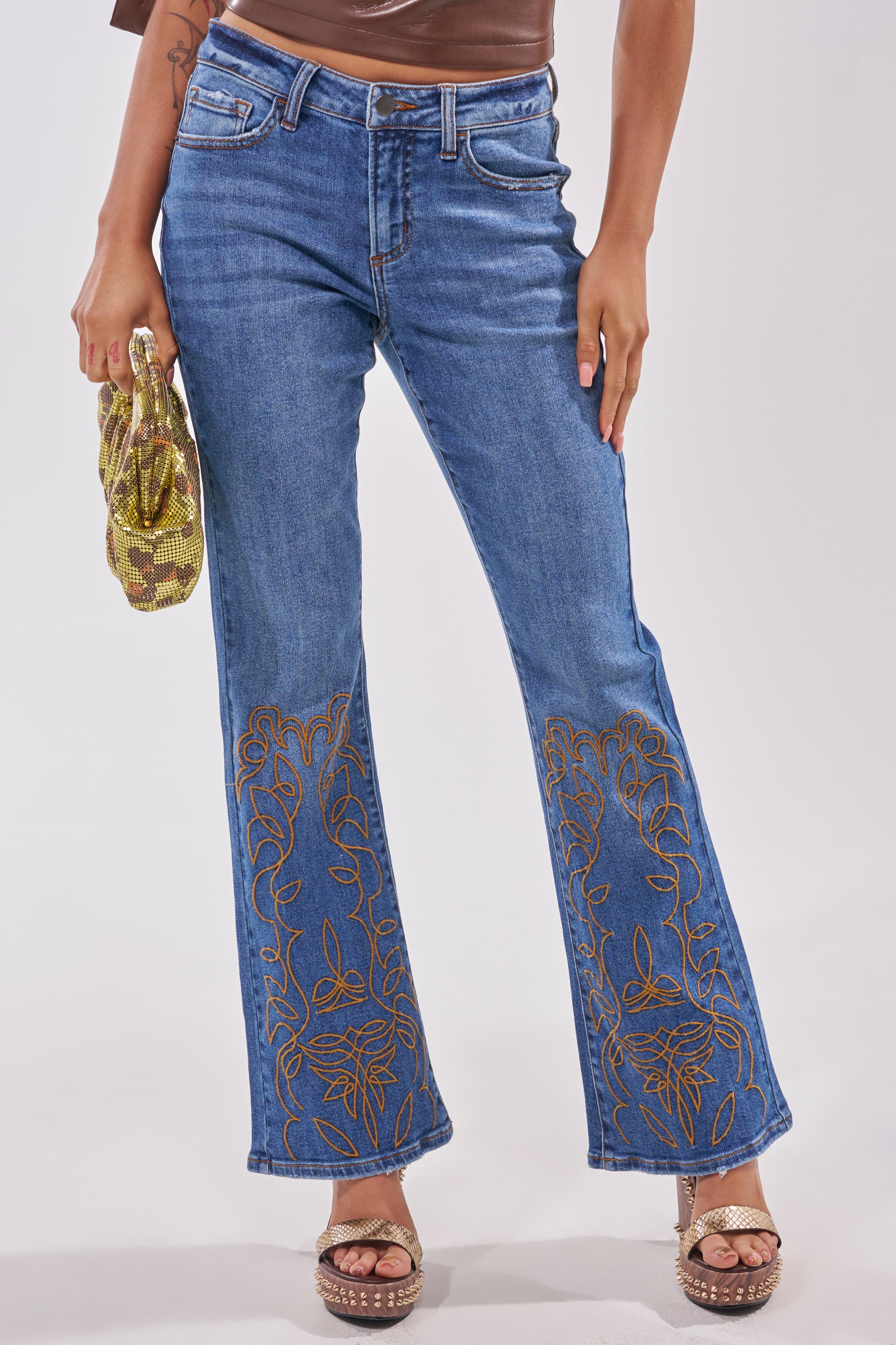 Wearing RODEO BABE SKINNY FLARED DENIM jeans with an embroidered hem, a person holds a gold sequin bag and stands in brown wedge sandals against a plain background. 1