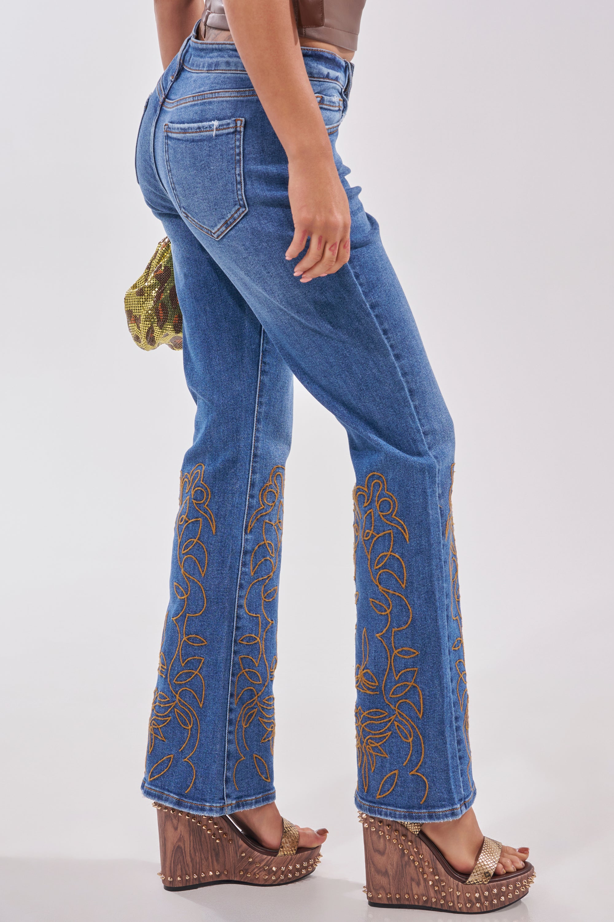 A person stands sideways against a plain background wearing the RODEO BABE SKINNY FLARED DENIM, tan platform wedge sandals, and holding a small green snakeskin handbag. 2