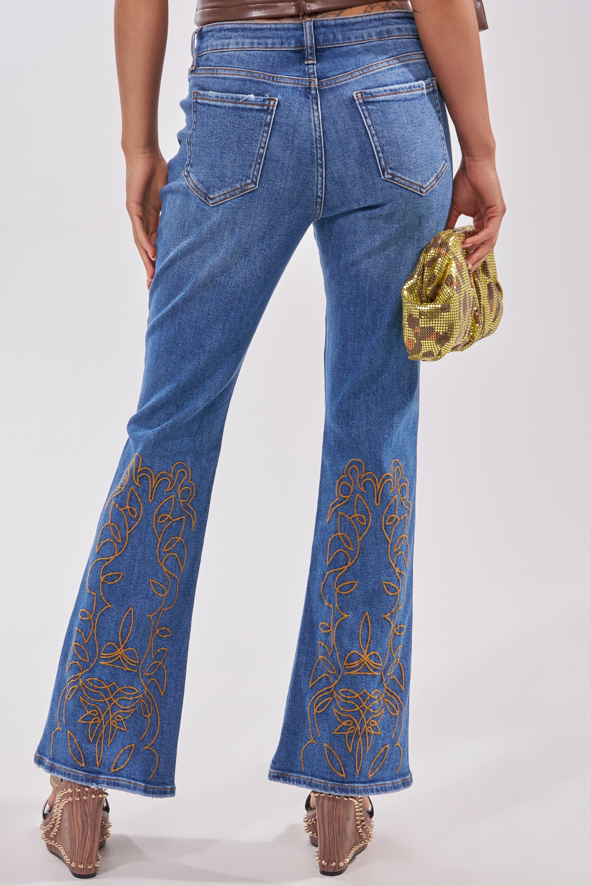 A person stands facing away, wearing RODEO BABE SKINNY FLARED DENIM with tan floral embroidery on the hem, brown heels, and holding a small gold sequined clutch. 3
