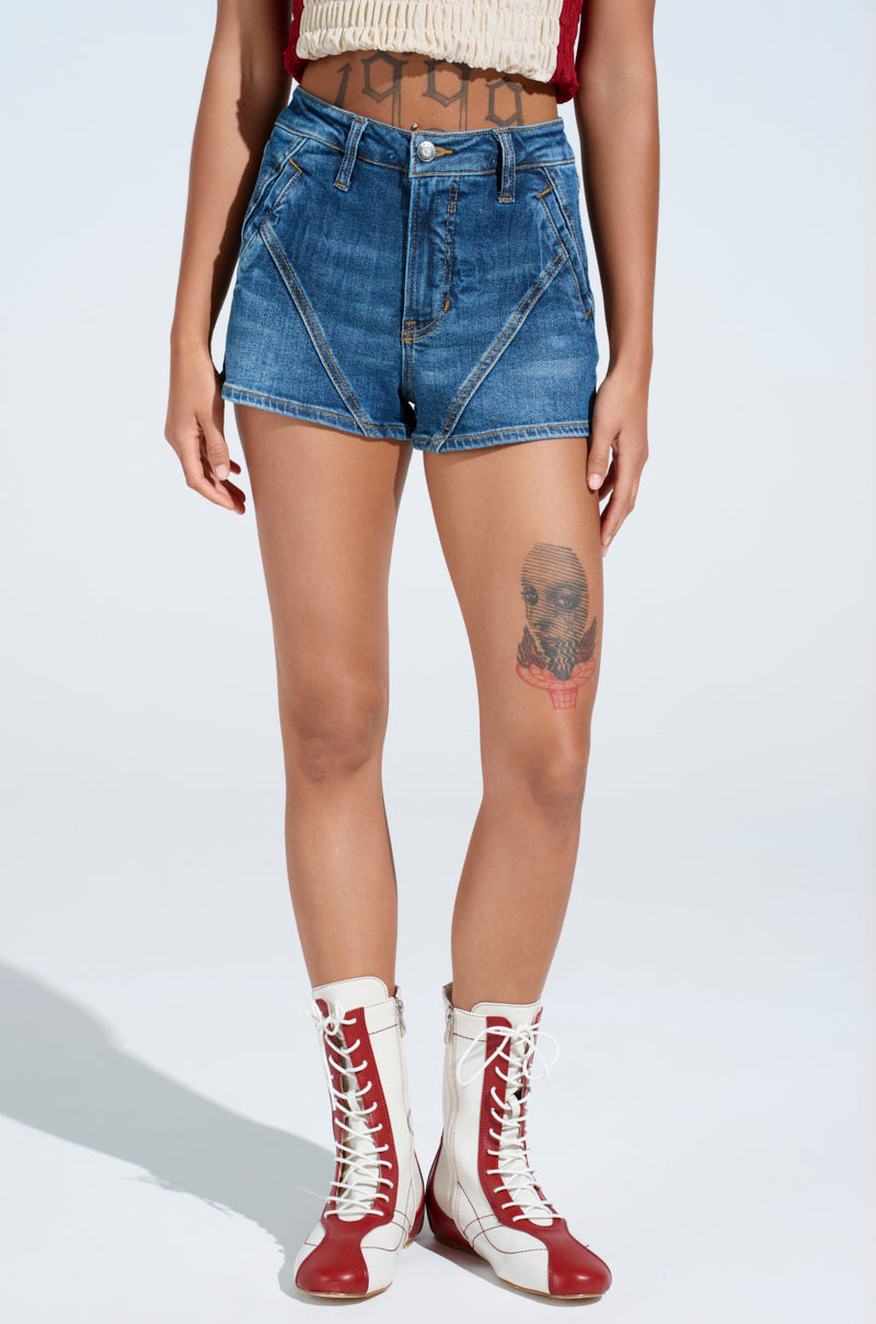 Wearing high-waisted LANA DENIM SHORT, a summer staple, paired with a crop top and red and white lace-up boots, they show off a thigh tattoo against a well-lit, plain backdrop. 2