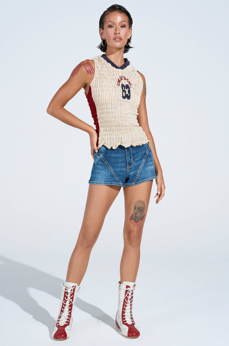 Confidently posed against a plain backdrop, a person wears a sleeveless cream top featuring a "B" and number design, the summer-essential LANA DENIM SHORT, and red and white high-top boots. They have short hair with visible leg and arm tattoos. 6