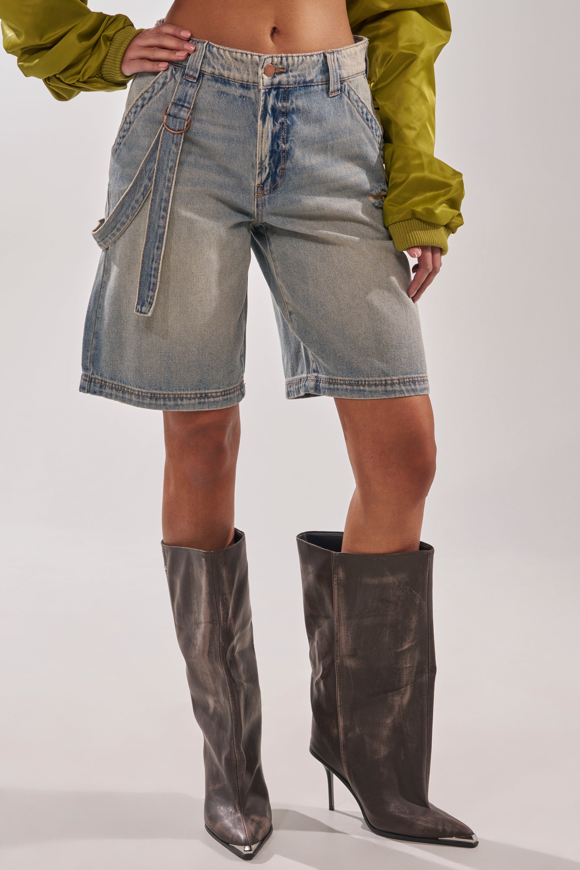A person wears the BABY ITS YOU DENIM SHORT, which are belted, knee-length bermuda shorts with a low-rise waist, paired with a green long-sleeve top and metallic brown pointed-toe knee-high boots. The image is cropped above the shoulders. 2