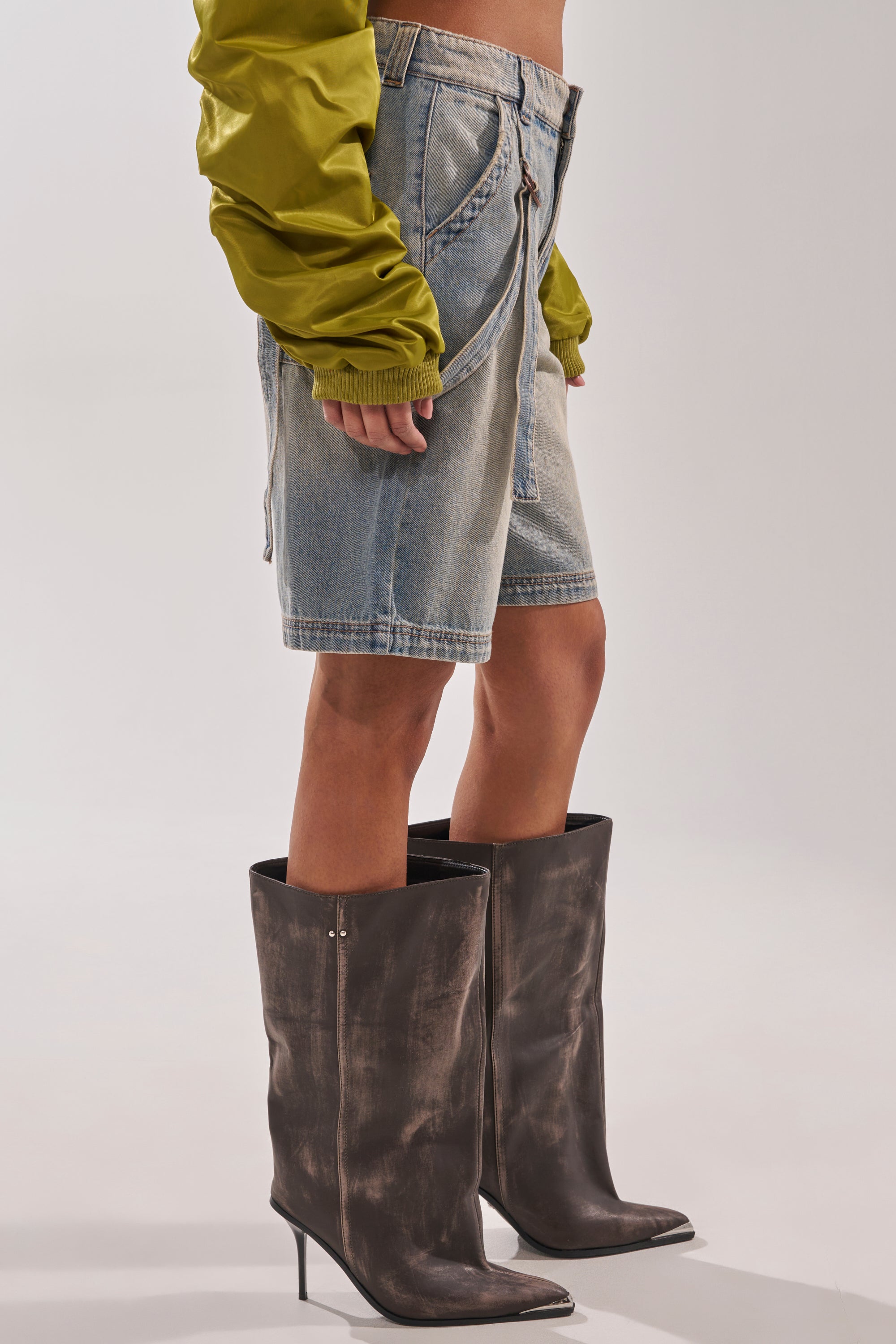 Wearing the BABY ITS YOU DENIM SHORT, a green long-sleeve top, and tall weathered black high-heeled boots, a person stands against a plain light background. 4