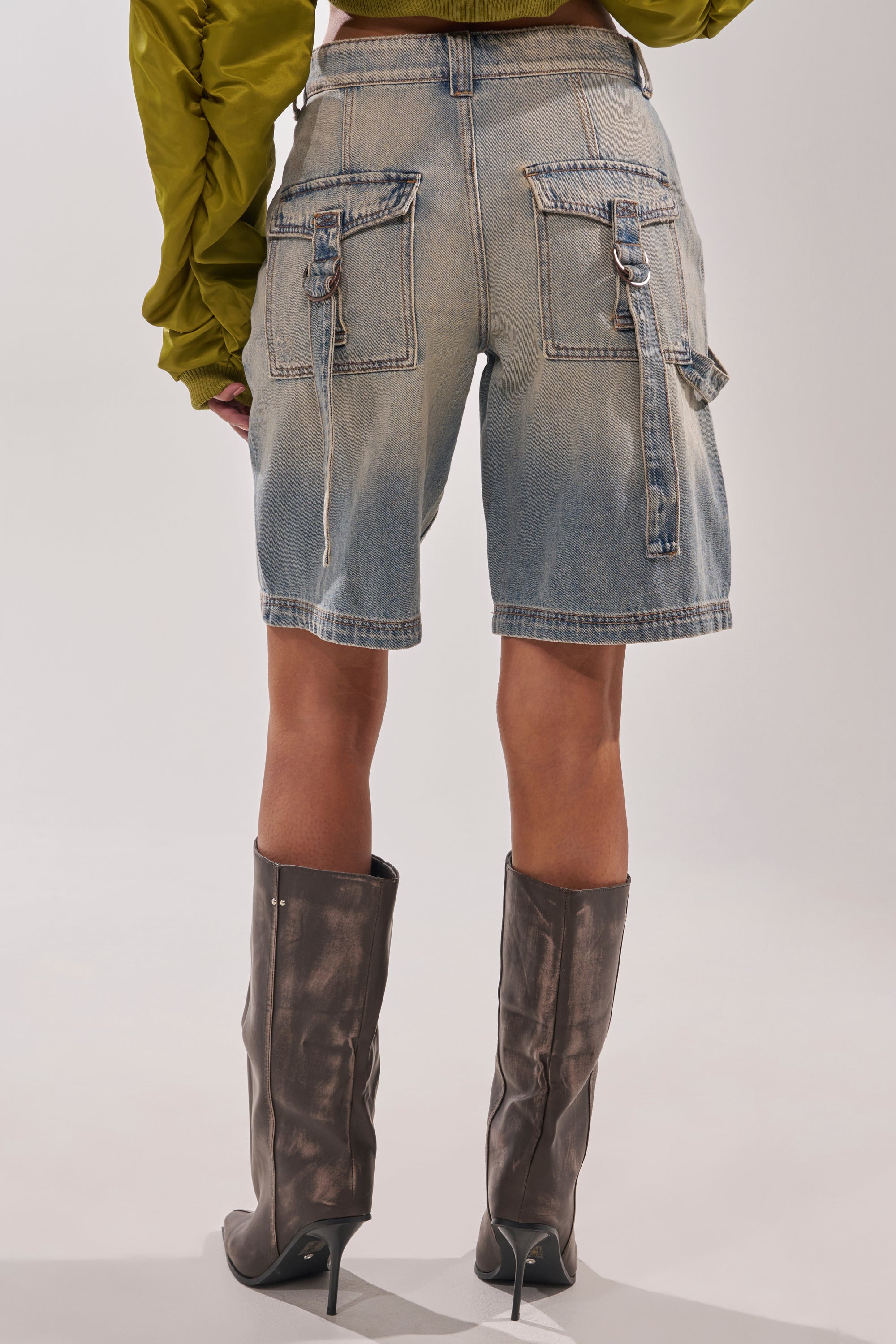 Model seen from the back wearing BABY ITS YOU DENIM SHORTS—knee-length, light-wash denim with a low rise, large back pockets, and decorative straps—paired with a green long-sleeve top and metallic heeled boots. 5