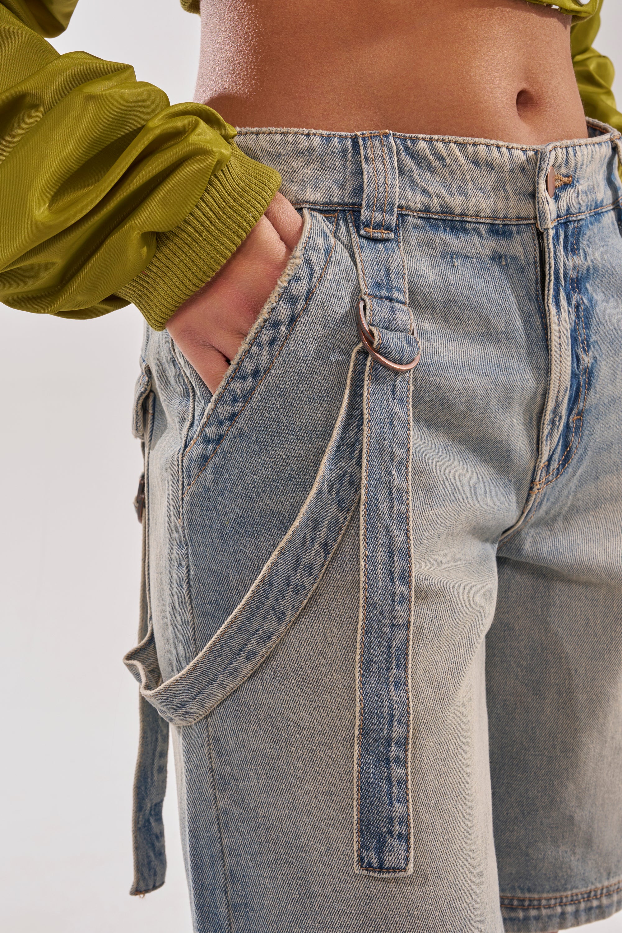 Wearing the BABY ITS YOU DENIM SHORT, a person sports high-waisted, light-wash denim with strap detail and a green cropped jacket. Their hand rests in the front pocket. Only the torso and thighs are shown; style contrasts current low-rise and bermuda trends. 7