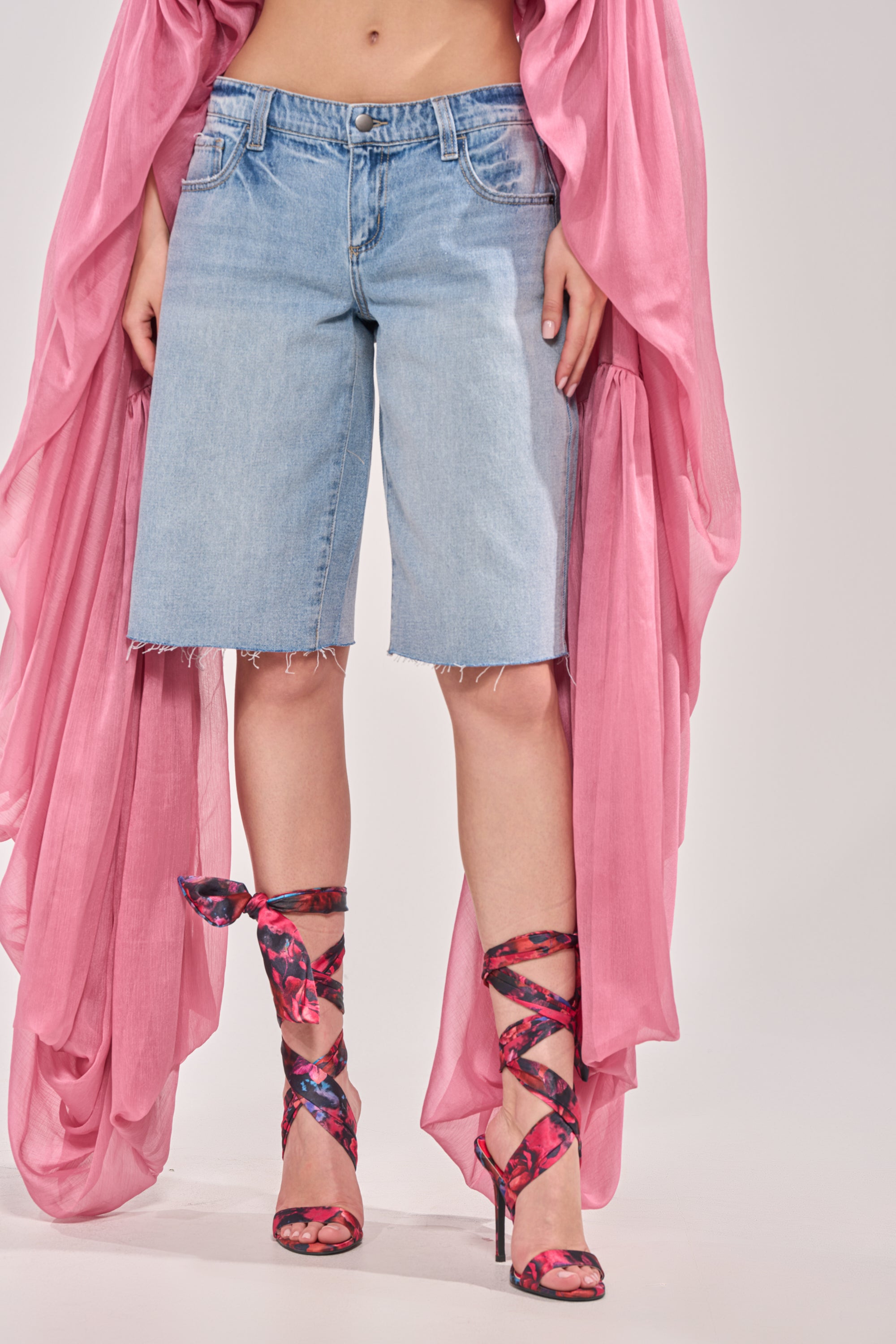 A person wears the ULTRA LOW RISE DENIM BERMUDA SHORT with a flowing pink sheer top and pink and purple wraparound high-heeled sandals, standing against a plain background. 0