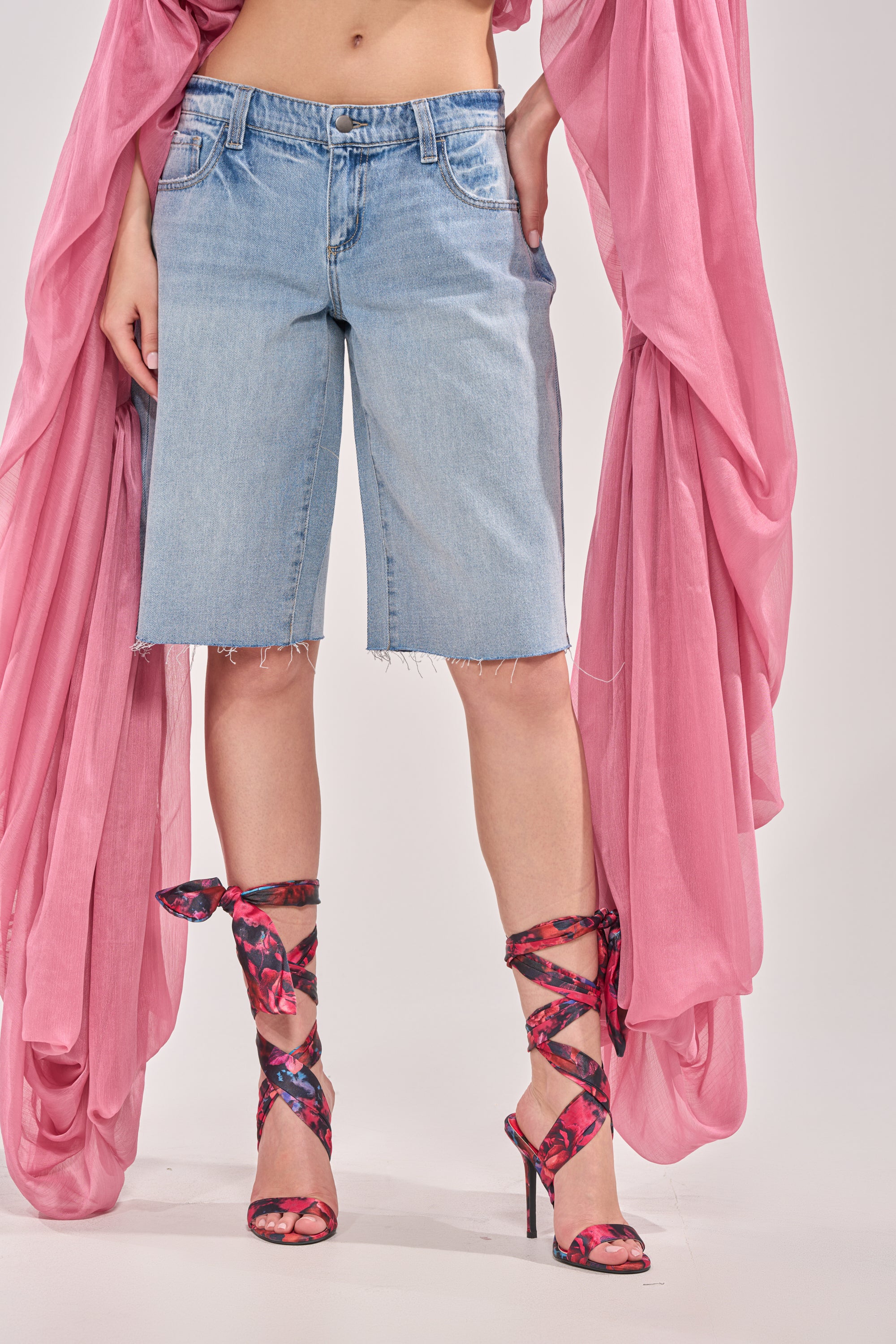 A person wears the ULTRA LOW RISE DENIM BERMUDA SHORTS, paired with a flowing pink sheer layer and pink-and-black strappy high heels. The image is cropped from the waist down, set against a plain background. 1