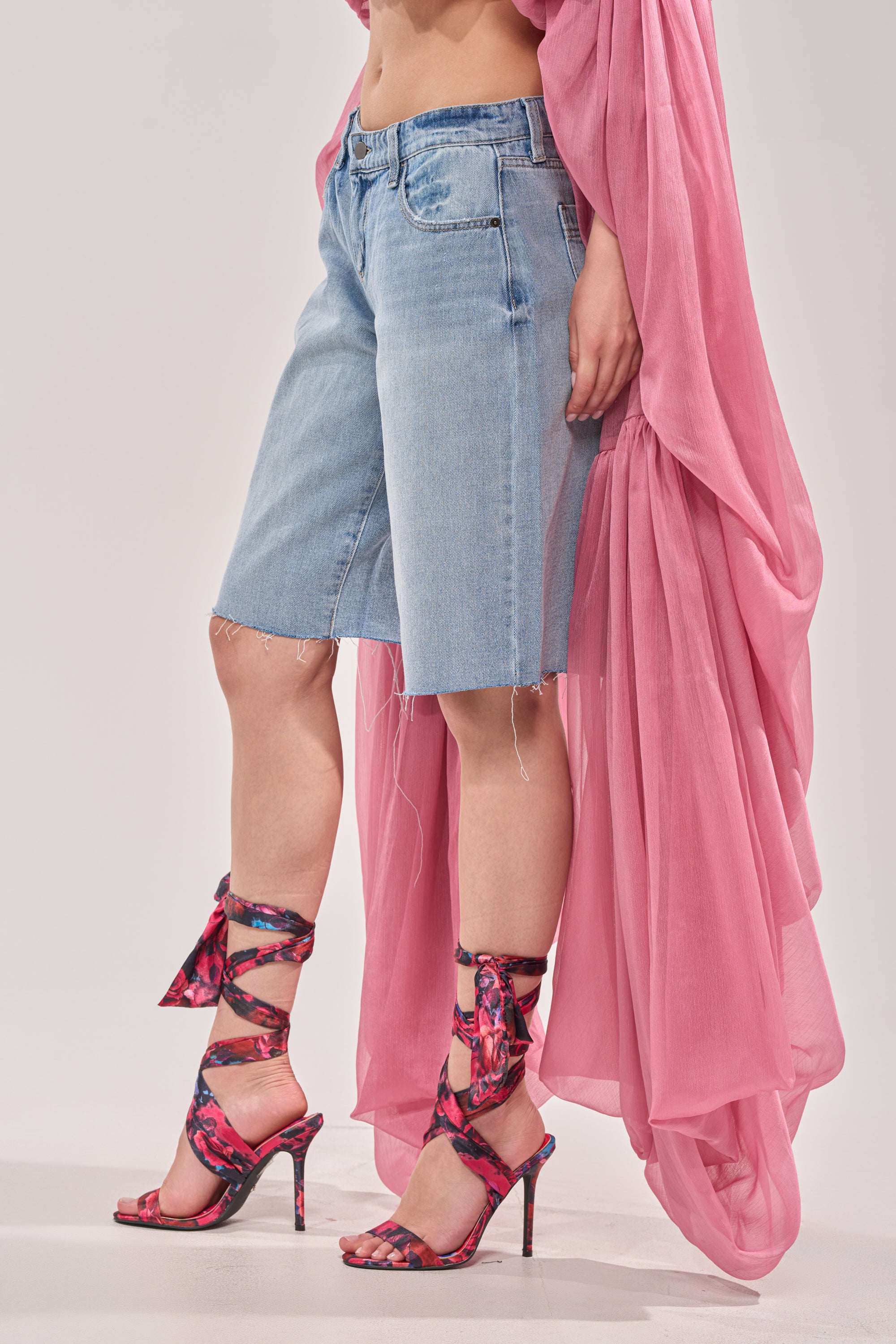 A person wears the ULTRA LOW RISE DENIM BERMUDA SHORT, a sheer pink top, and floral pink strappy high heels, posing against a light background. 2