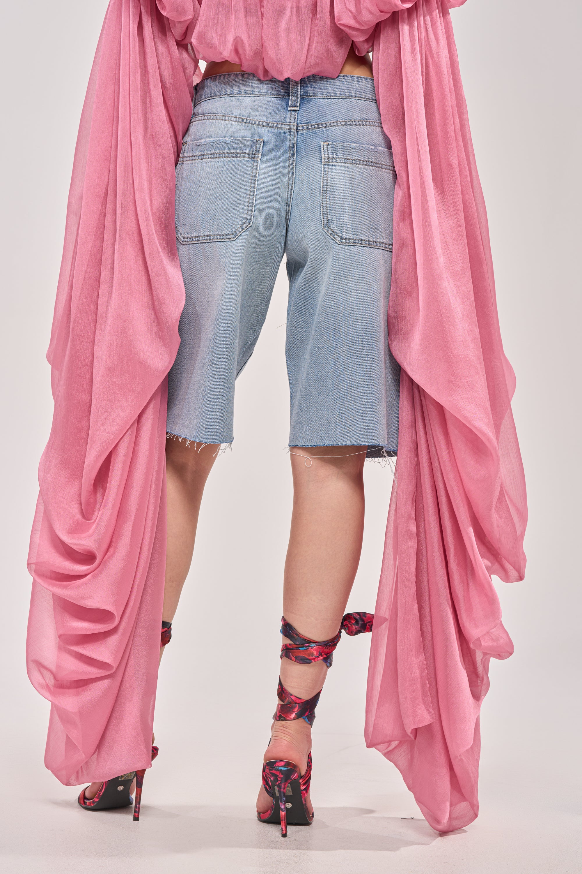 A person is seen from behind wearing ULTRA LOW RISE DENIM BERMUDA SHORTS, a pink flowing top with long draped fabric, and matching pink floral high heels with ribbon ties against a plain background. 3
