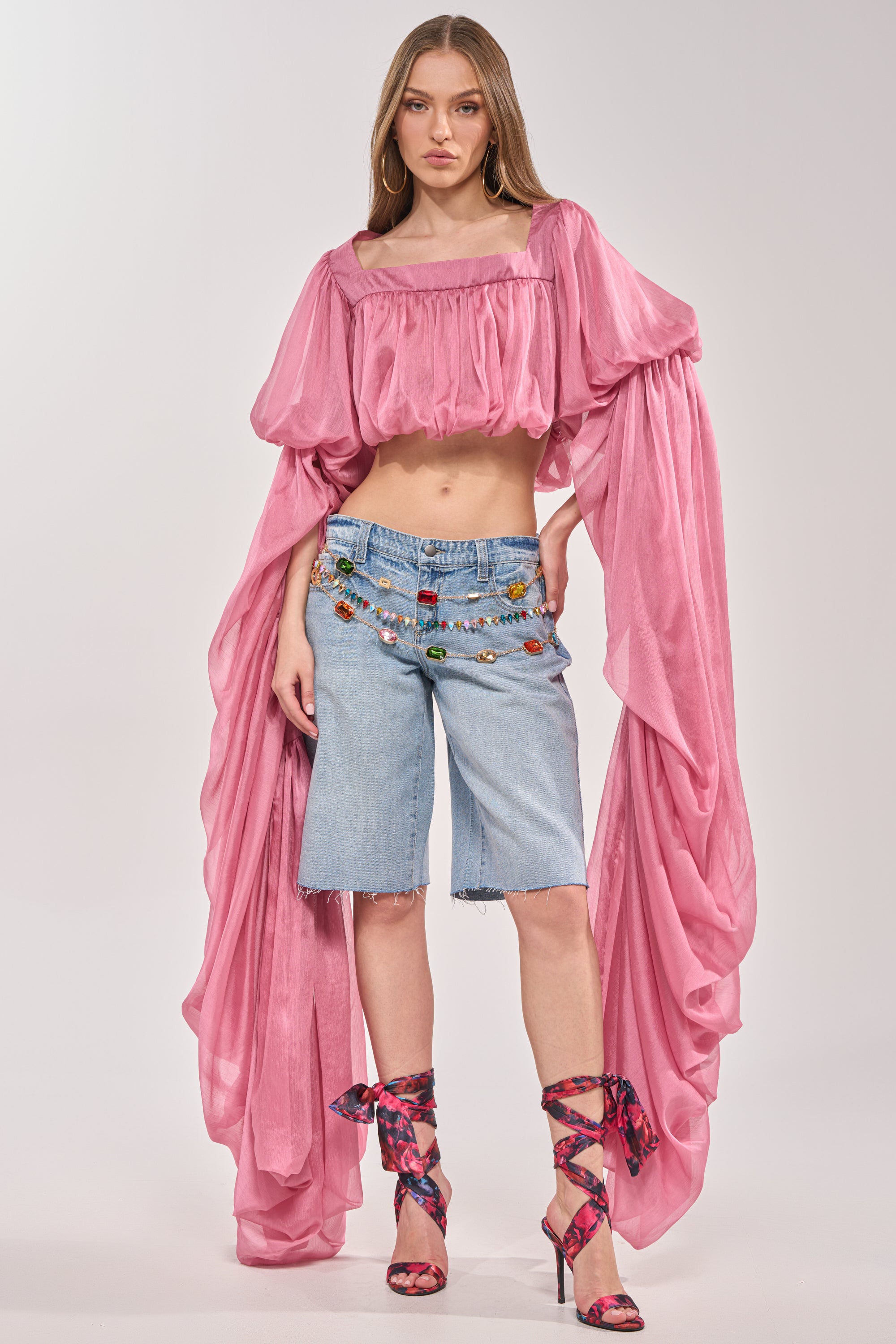 A woman stands confidently in a flowing pink off-shoulder top, a colorful beaded belt, matching pink patterned high-heeled sandals tied at the ankle, and ULTRA LOW RISE DENIM BERMUDA SHORT. 4