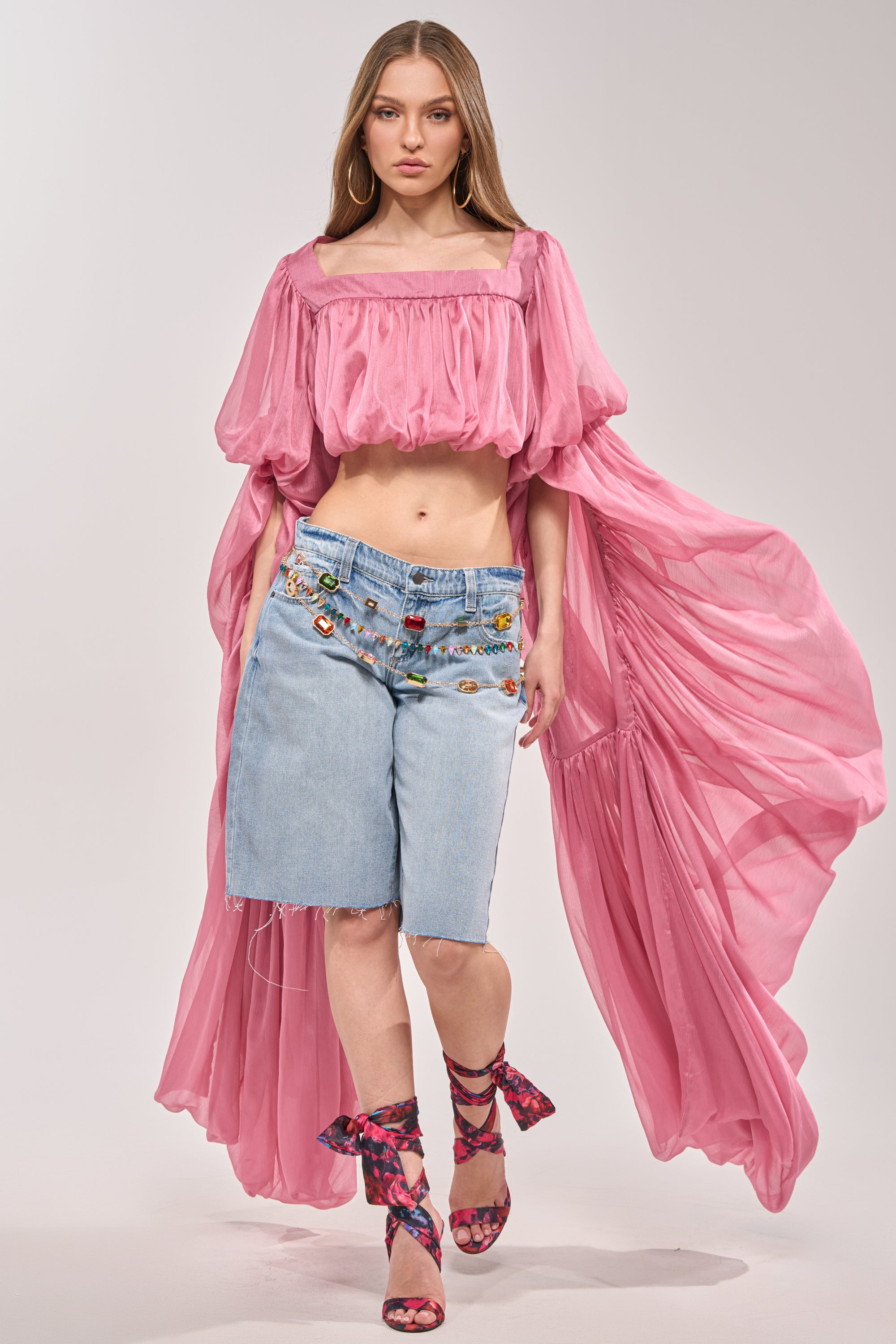 A woman wears ULTRA LOW RISE DENIM BERMUDA SHORT paired with a sheer pink off-shoulder blouse and matching pink high-heel sandals, posing against a plain background. 5