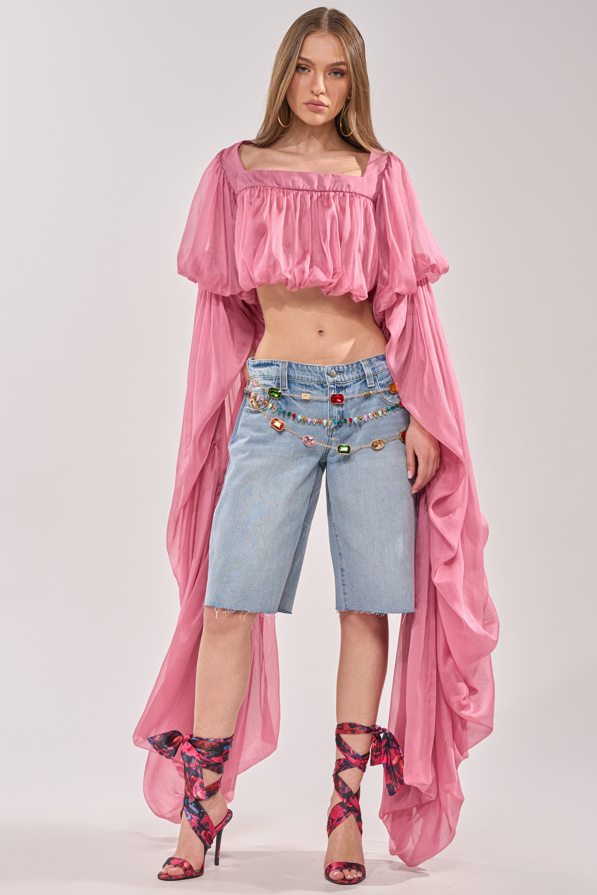 A woman poses in a dramatic pink off-shoulder crop top, a colorful beaded belt, matching pink lace-up heels, and ULTRA LOW RISE DENIM BERMUDA SHORT against a plain background. 6