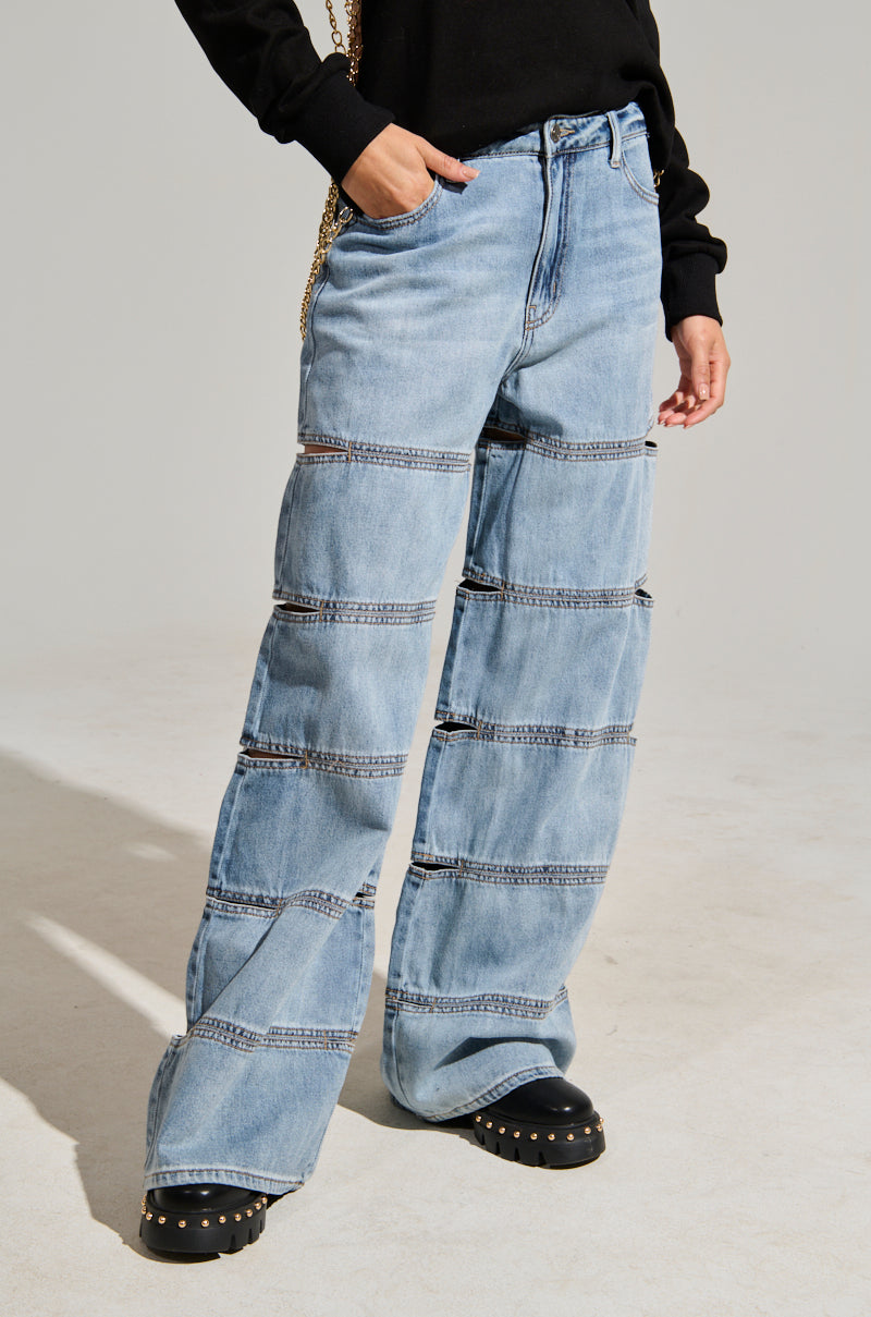Dressed in the WE GROOVIN DENIM PANT, featuring its light blue color and high-rise fit with horizontal seams, a person pairs it with a black sweater and casually rests their hand in the jeans' pocket. Their ensemble is completed with black shoes adorned with studded details. A plain light gray background highlights the wide-leg silhouette of their look.