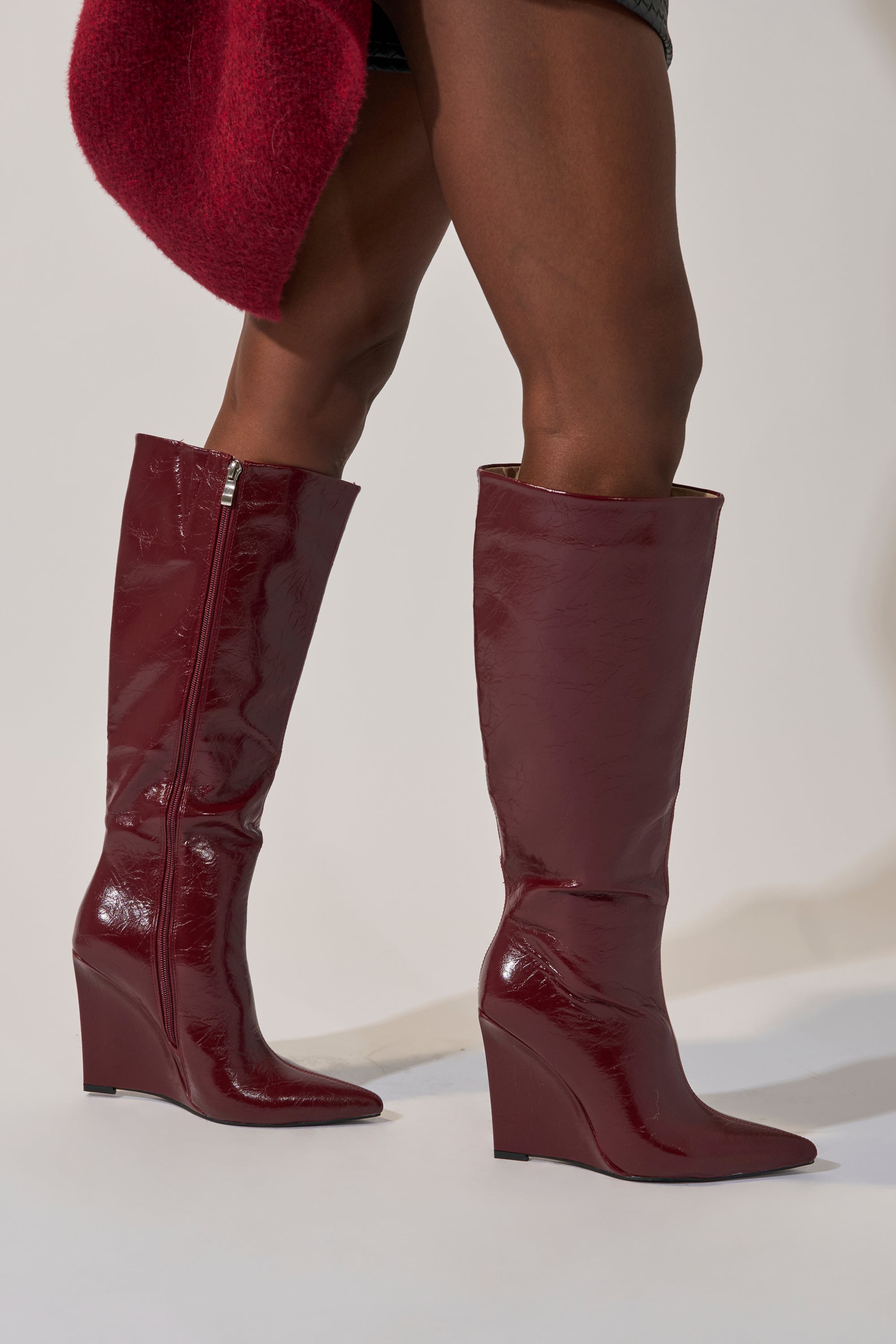 A person wearing the AZALEA WANG AMAADIS BURGUNDY WEDGE KNEE HIGH BOOT, featuring shiny wedge heels, and a textured red garment, stands on a light background.
