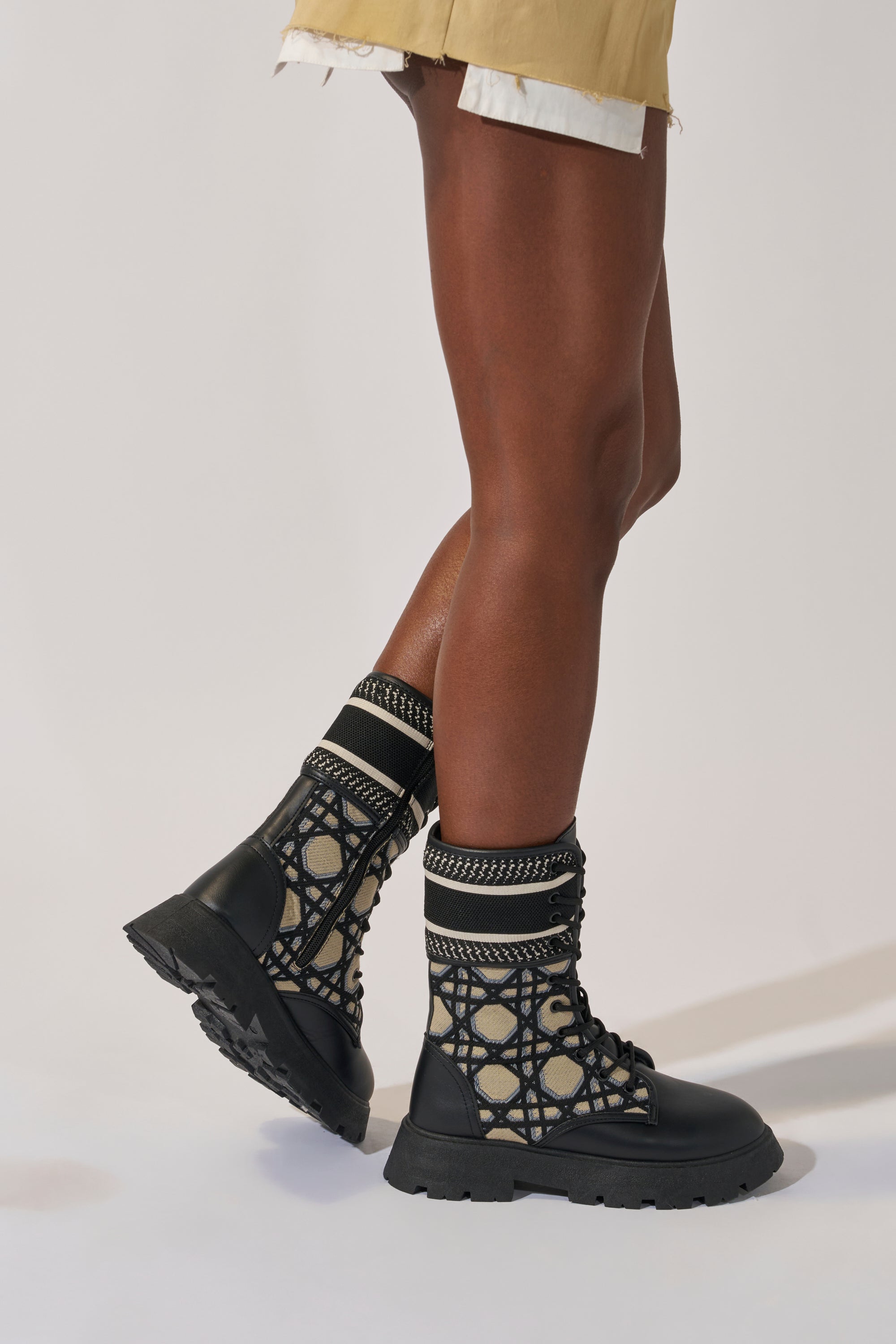 A person in beige shorts wears AMADA CANVAS PRINT COMBAT BOOT—black lace-up combat boots with a mesh design and thick soles—standing on a light background, one leg crossed for a modern streetwear vibe.