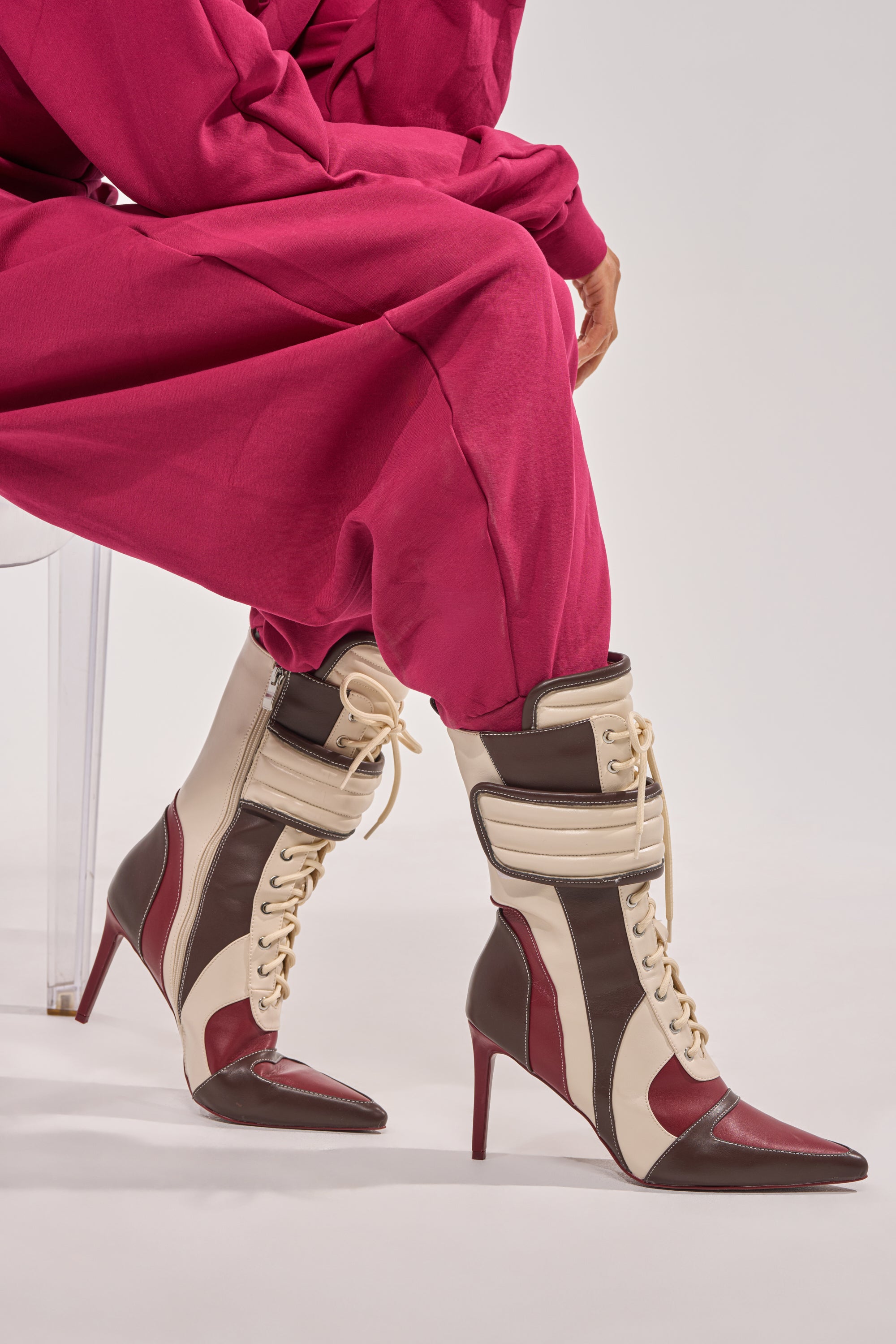 A person in magenta pants sits on a clear chair, wearing the AZALEA WANG AMADA BROWN SPORTY STILETTO BOOTIE featuring a pointed toe and lace-up design. Only the lower half of the body is visible.