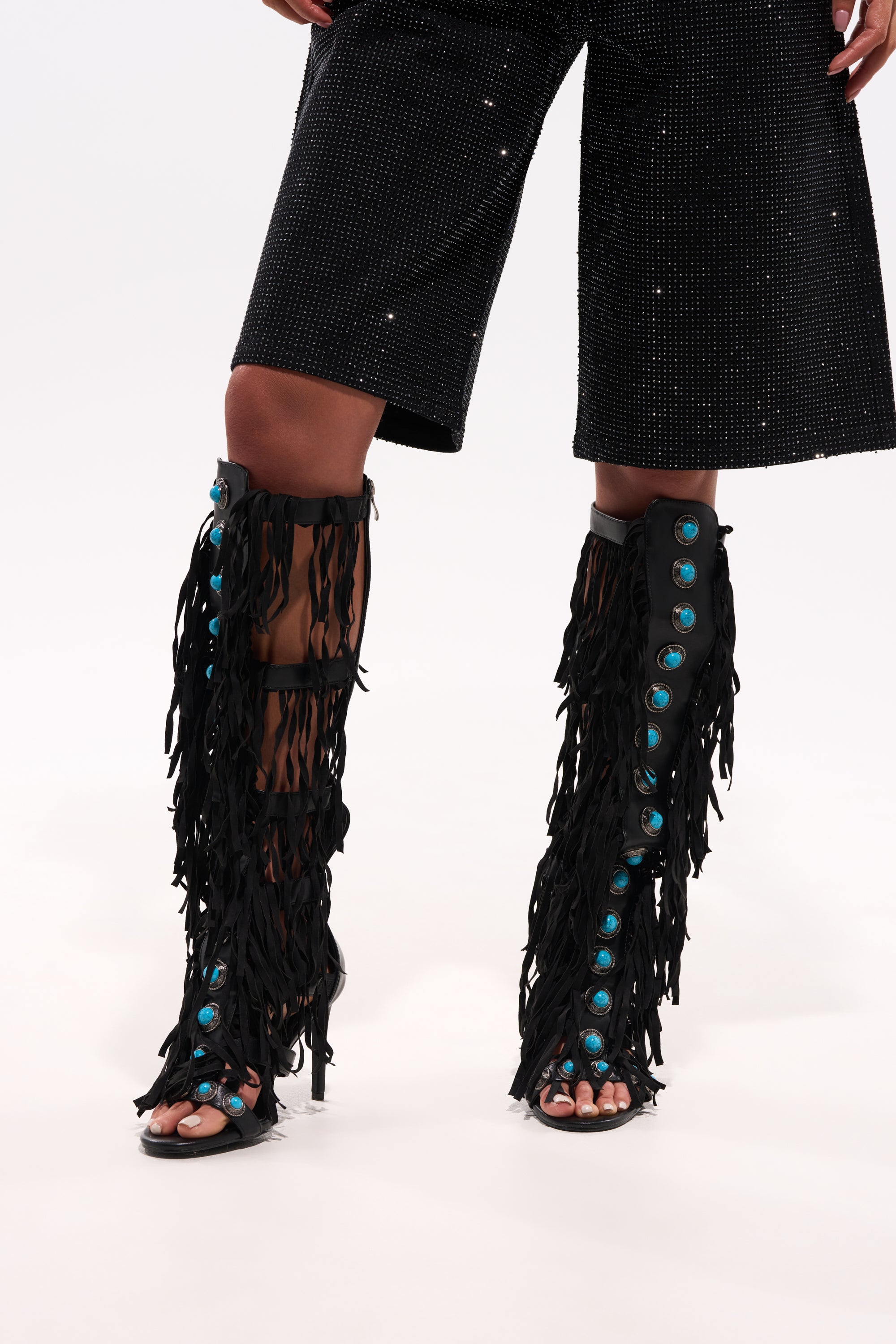 Someone stands against a white background wearing black sequined knee-length shorts and the AZALEA WANG AMADEO BLACK FRINGE AND TURQUOISE GLADIATOR STILETTO SANDAL. 0