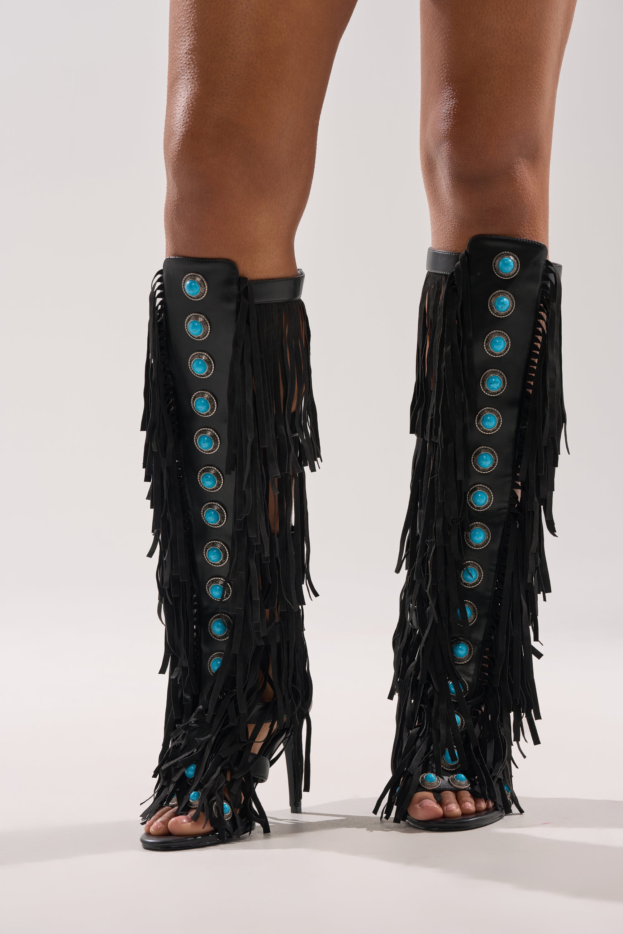 A person models the AZALEA WANG AMADEO Black Fringe and Turquoise Gladiator Stiletto Sandal, featuring black fringe and turquoise accents, while standing on a light gray background. 1
