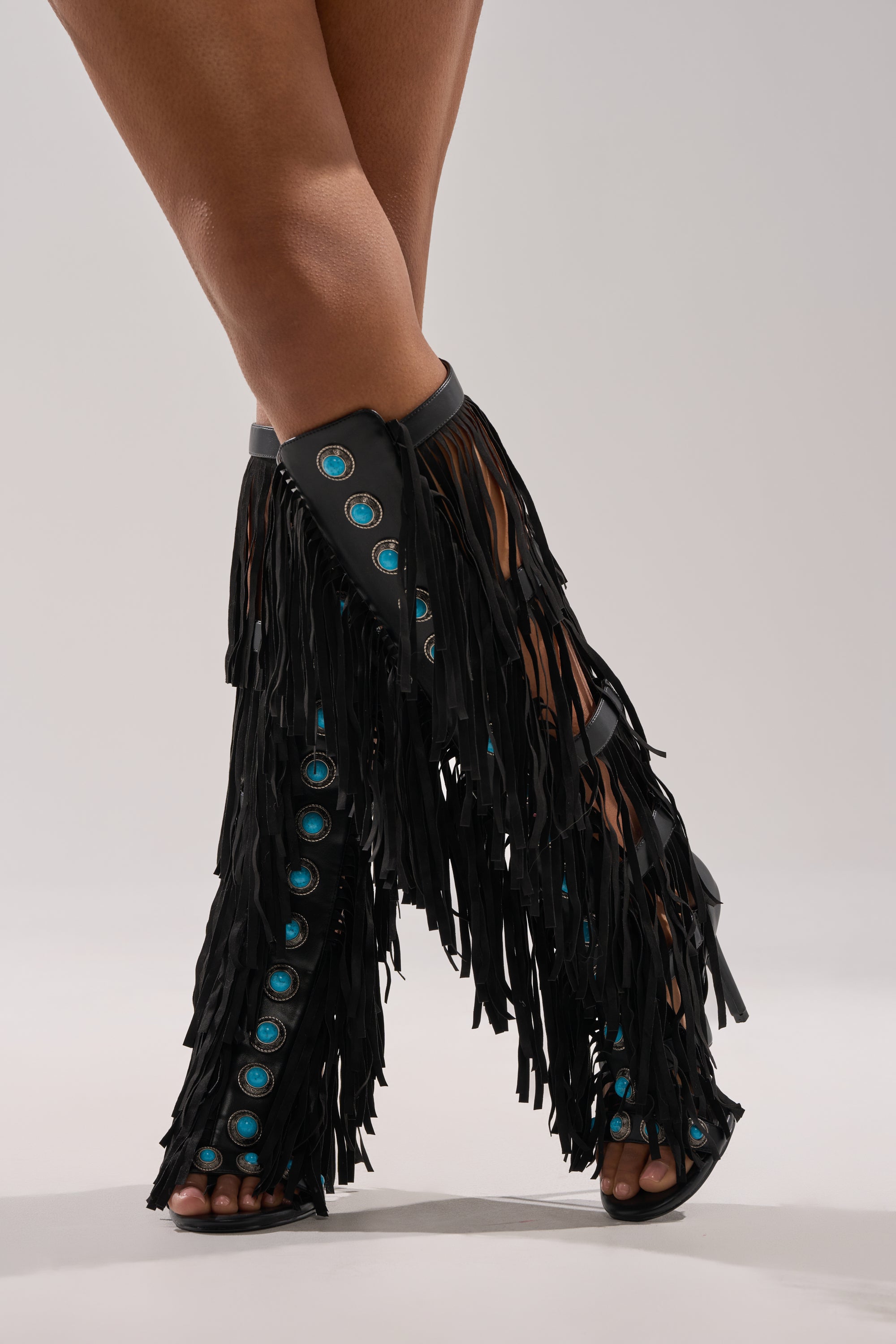 A person stands with legs crossed against a neutral background, wearing the AZALEA WANG AMADEO BLACK FRINGE AND TURQUOISE GLADIATOR STILETTO SANDAL. 3