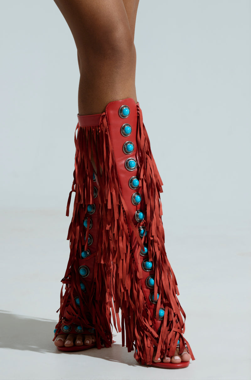 Against a plain background, a person wears the AZALEA WANG AMADEO RED SANDAL—red knee-high fringe boots with turquoise gemstone embellishments. These open-toed gladiator-style sandals offer a unique and stylish edge.