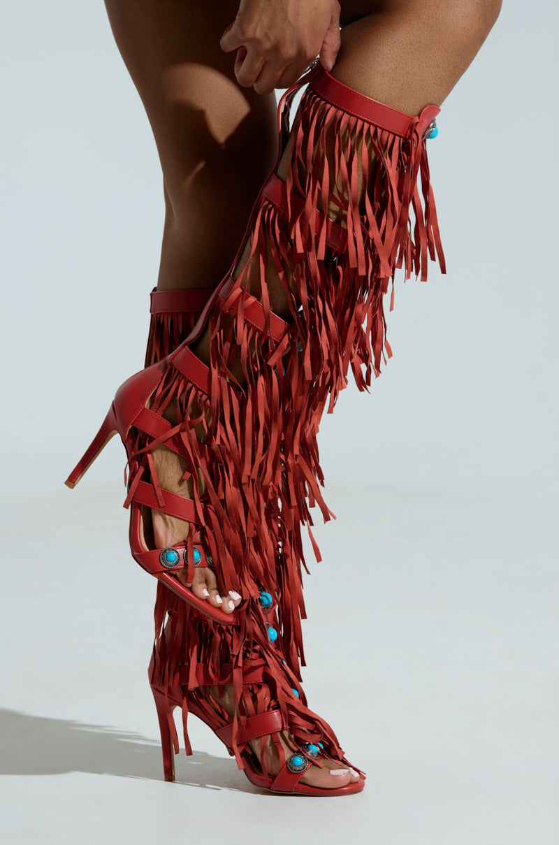 A person wears the AZALEA WANG AMADEO RED SANDAL, featuring a tall gladiator silhouette with red high heels, long fringe, turquoise accents, gemstone embellishments, multiple straps, and an open toe design. The background is plain light gray.